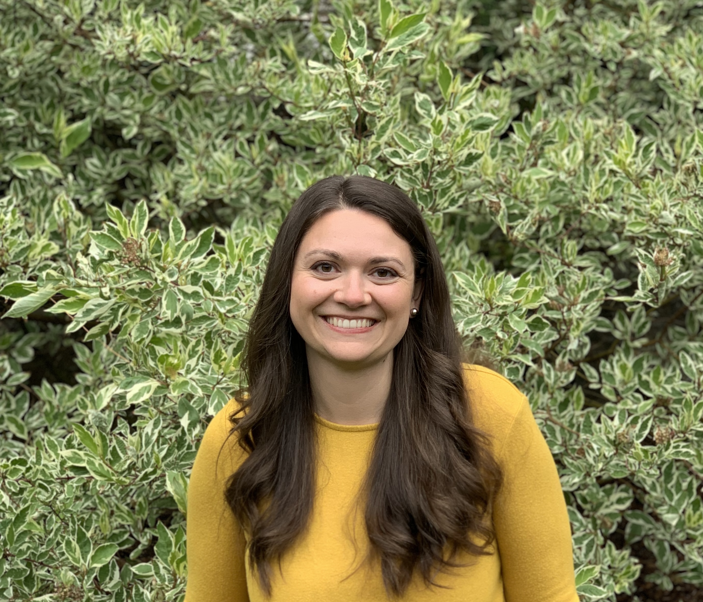 A smiling person with long brown hair in a yellow sweater stands in front of a hedge