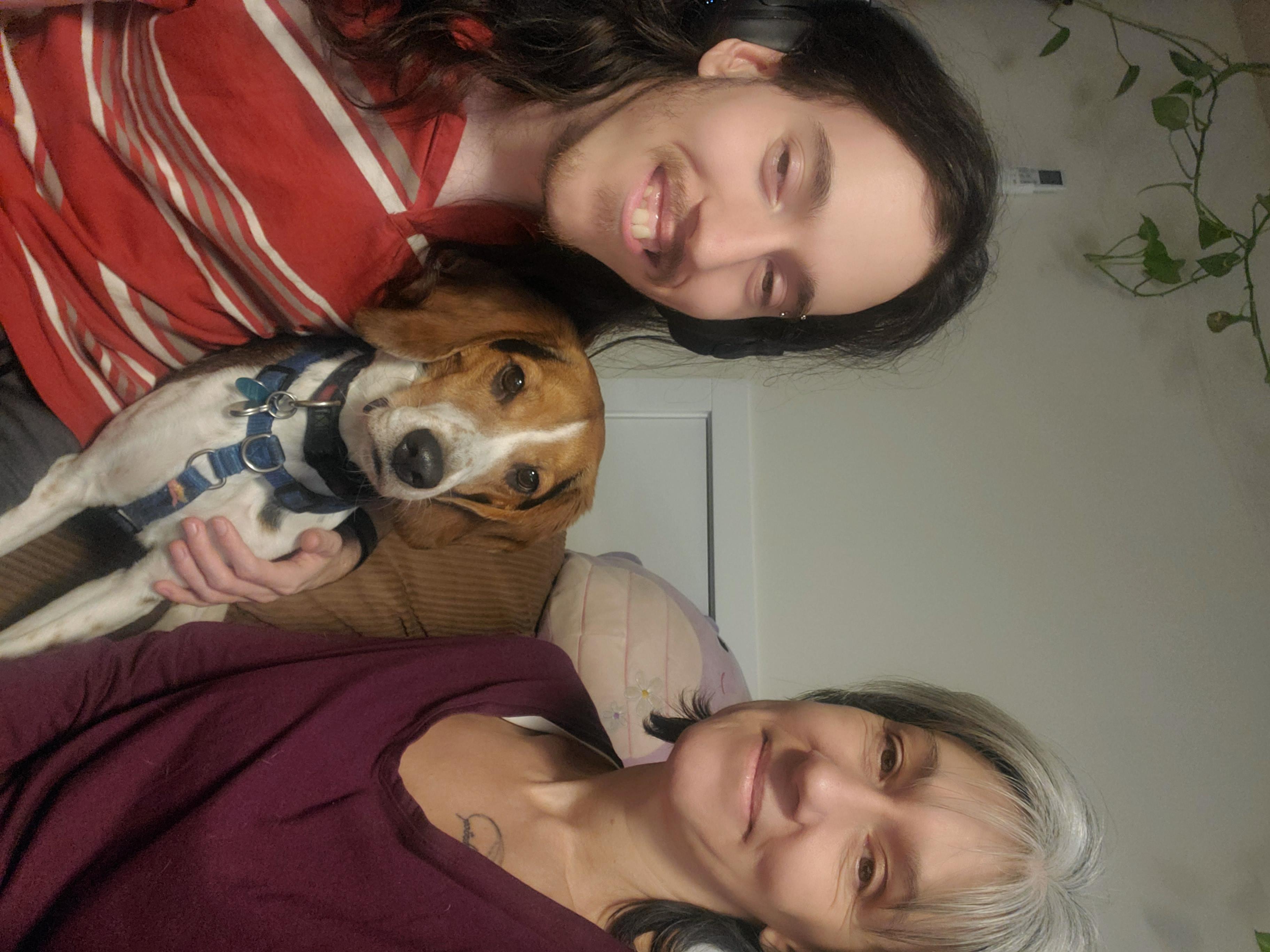 A young man with long brown hair wearing a red striped shirt holds a brown and white beagle next to a smiling woman with grey medium-length hair wearing a burgundy top