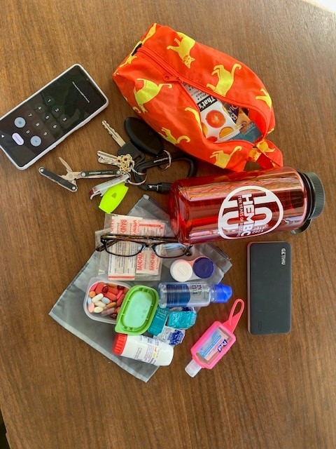 ed patterned toiletries case, red water bottle, cell phone, keys, glasses, medications, hand sanitizer, and other items lying on a table.