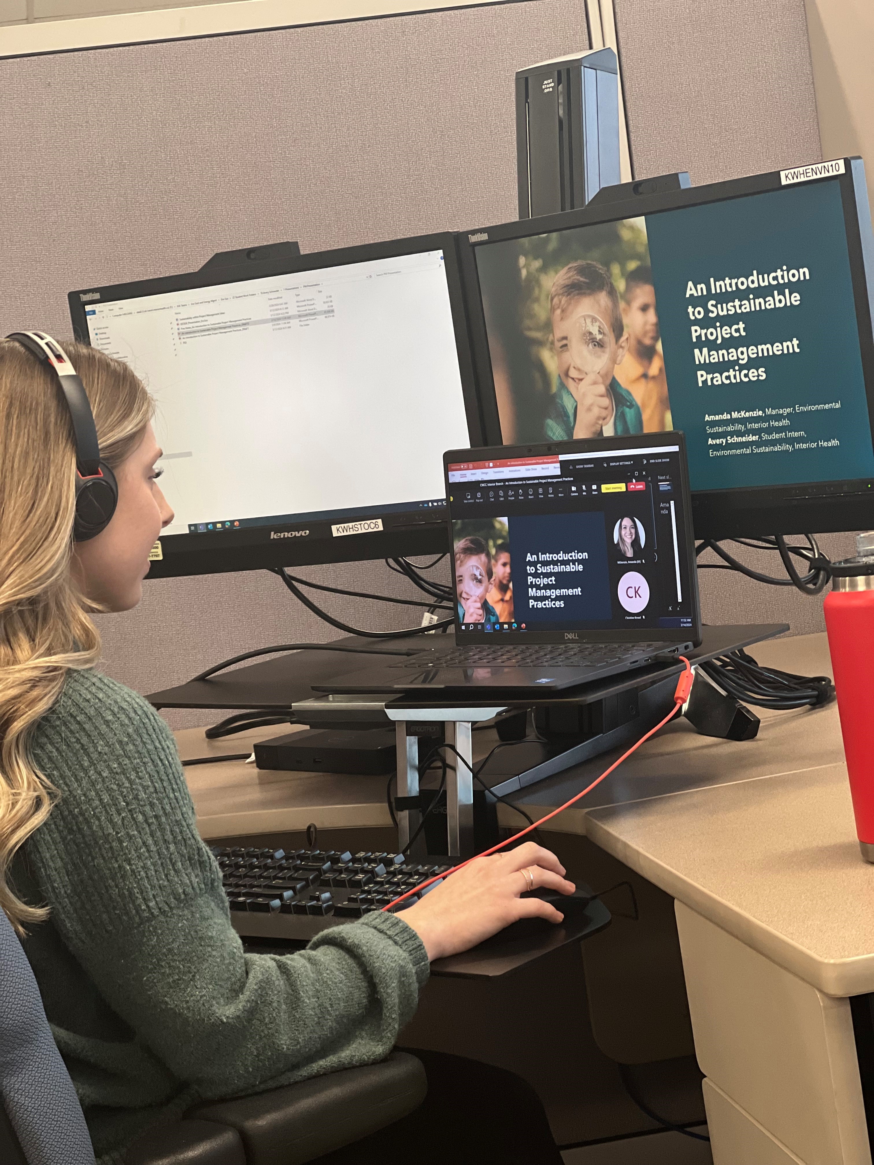 A young woman in a green sweater in front of two monitors and a laptop during an online presentation.