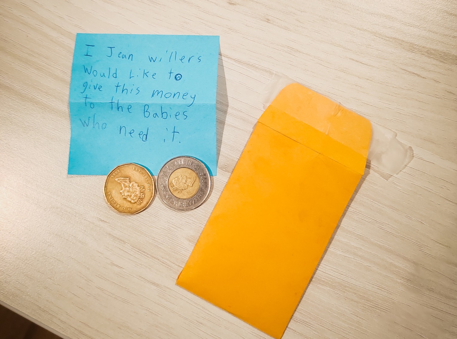 A small yellow manila envelope sits next to a toonie and a loonie sitting on top a blue note that says I Jean Willers would like to give this money to the babies who need it.