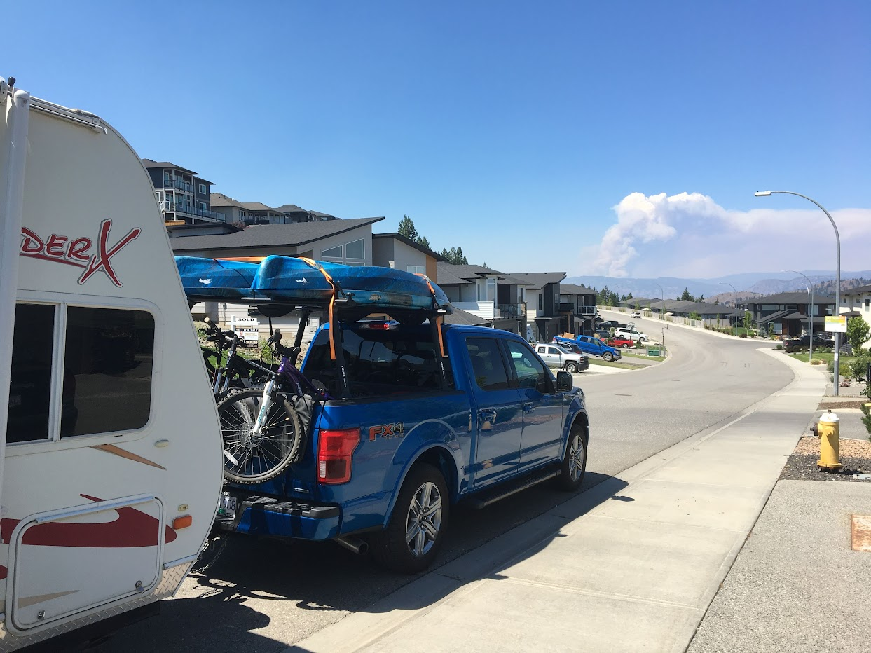 Blue truck parked on road loaded with kayaks and bikes pulling a trailer with a cloud of smoke in the distance.
