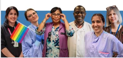 Group of five diverse healthcare professionals standing in a row, each wearing different styles of work attire including scrubs and lab coats. One individual is holding a small rainbow flag. They are smiling and appear in a friendly and professional setting.