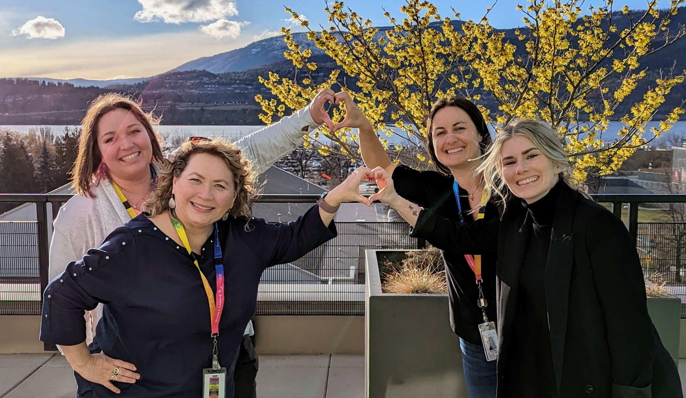 Four people smiling and posing outdoors on a balcony, forming a heart shape with their hands, with a scenic view of hills and a partly cloudy sky in the background.