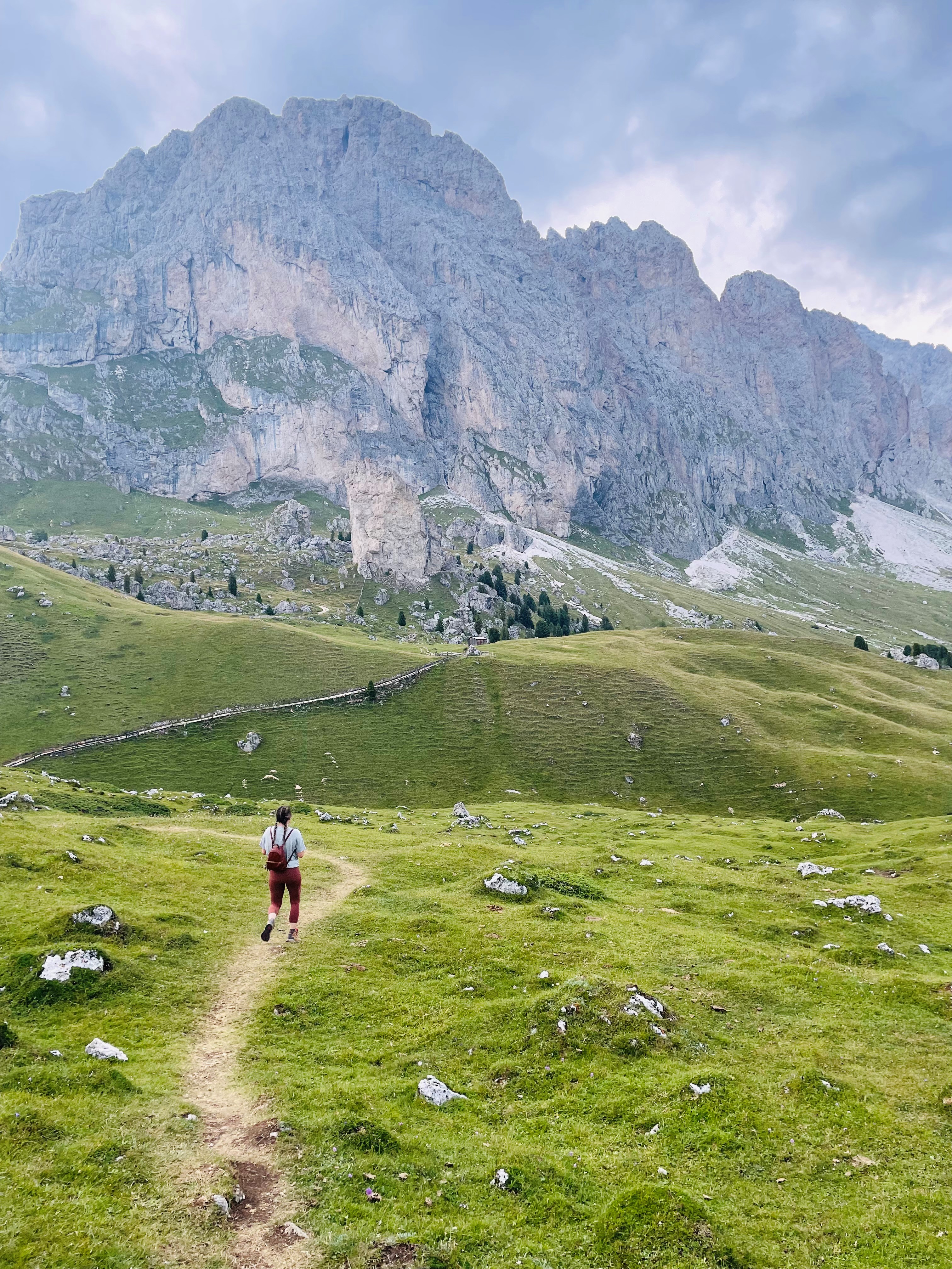 A photo a woman wearing a light blue tee shirt, red leggings and a red backpack walking towards the Dolomite Mountains, Dolomite Alps or Dolomitic Alps, a mountain range in northeastern Italy. 