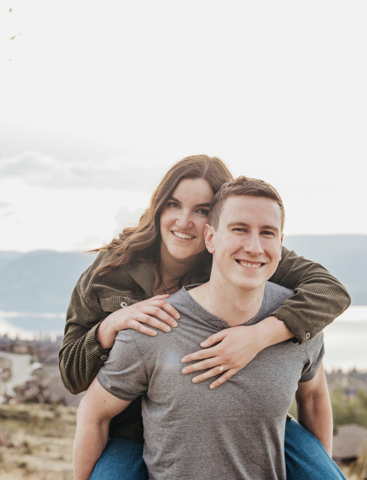 A woman with long brown hair in a brown corduroy shirt piggy backs on a man in a grey t-shirt