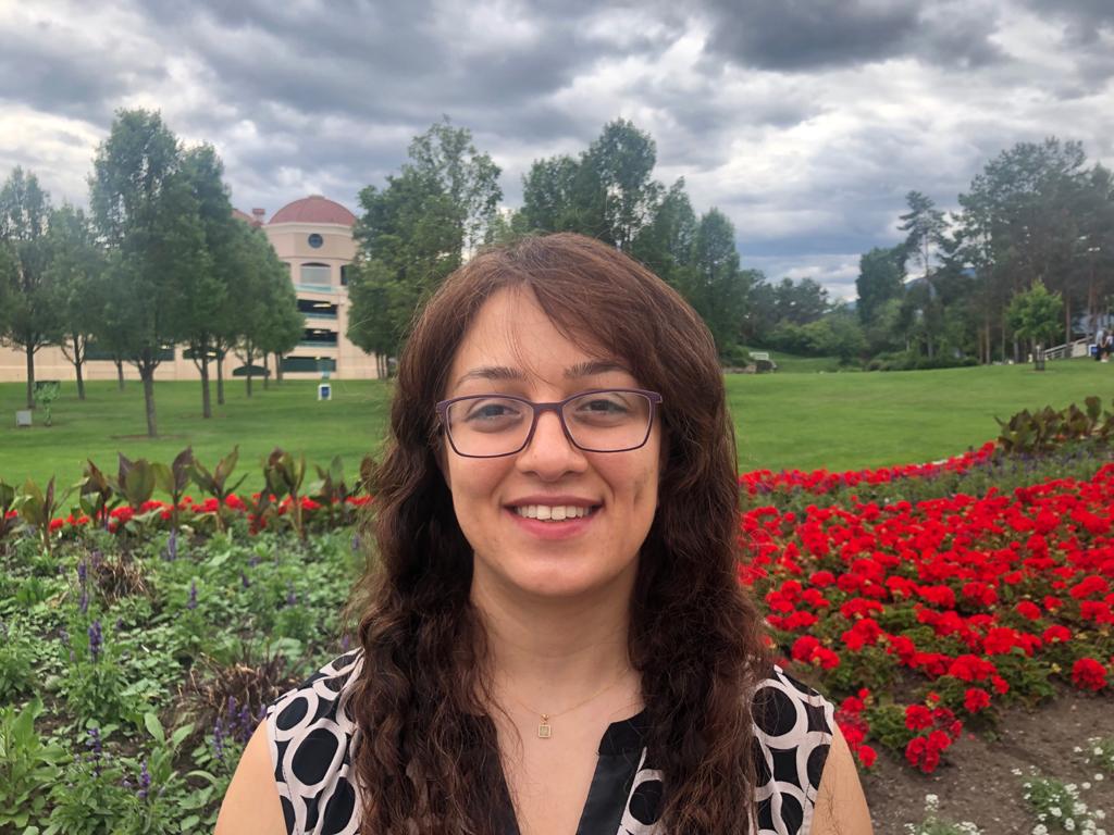 A woman with long red-brown hair and brown square glasses in a black and white sleeveless blouse stands outside in front of a garden of red flowers with trees and grass in the background.