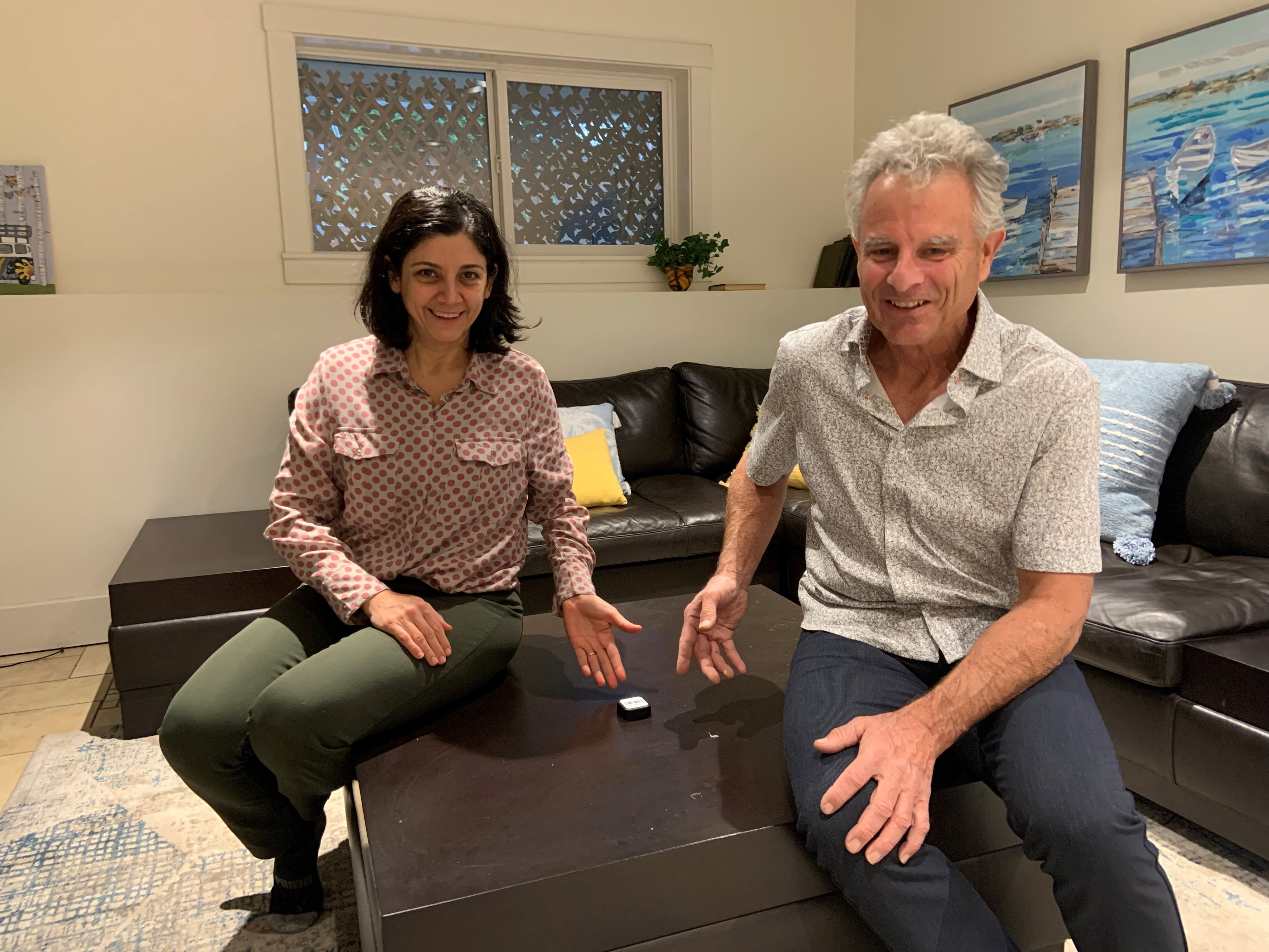 A woman with short black hair wearing a pink polka-dot shirt and green pants and a man with a grey short-sleeved shirt and blue pants sit together on an ottoman pointing at a small white square, a radon detector.