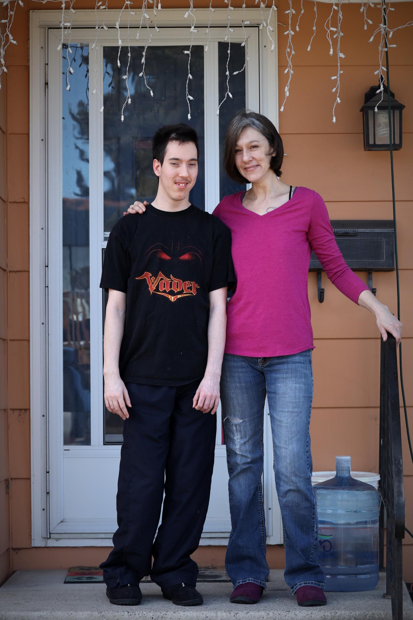 A young man with short dark hair wearing a black t-shirt and pants stands next to a woman with short dark grey hair wearing a pink long-sleeved shirt and blue jeans on a porch with a white door and white lights overhead.