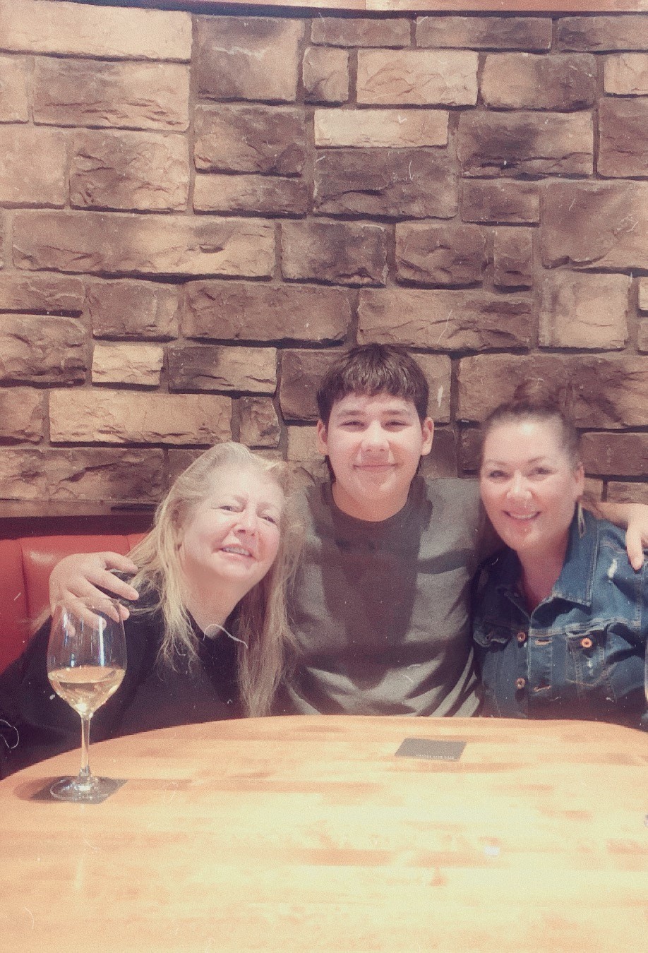 Three people smiling and posing at a table in front of a stone wall backdrop, with a glass of wine and a menu on the table.