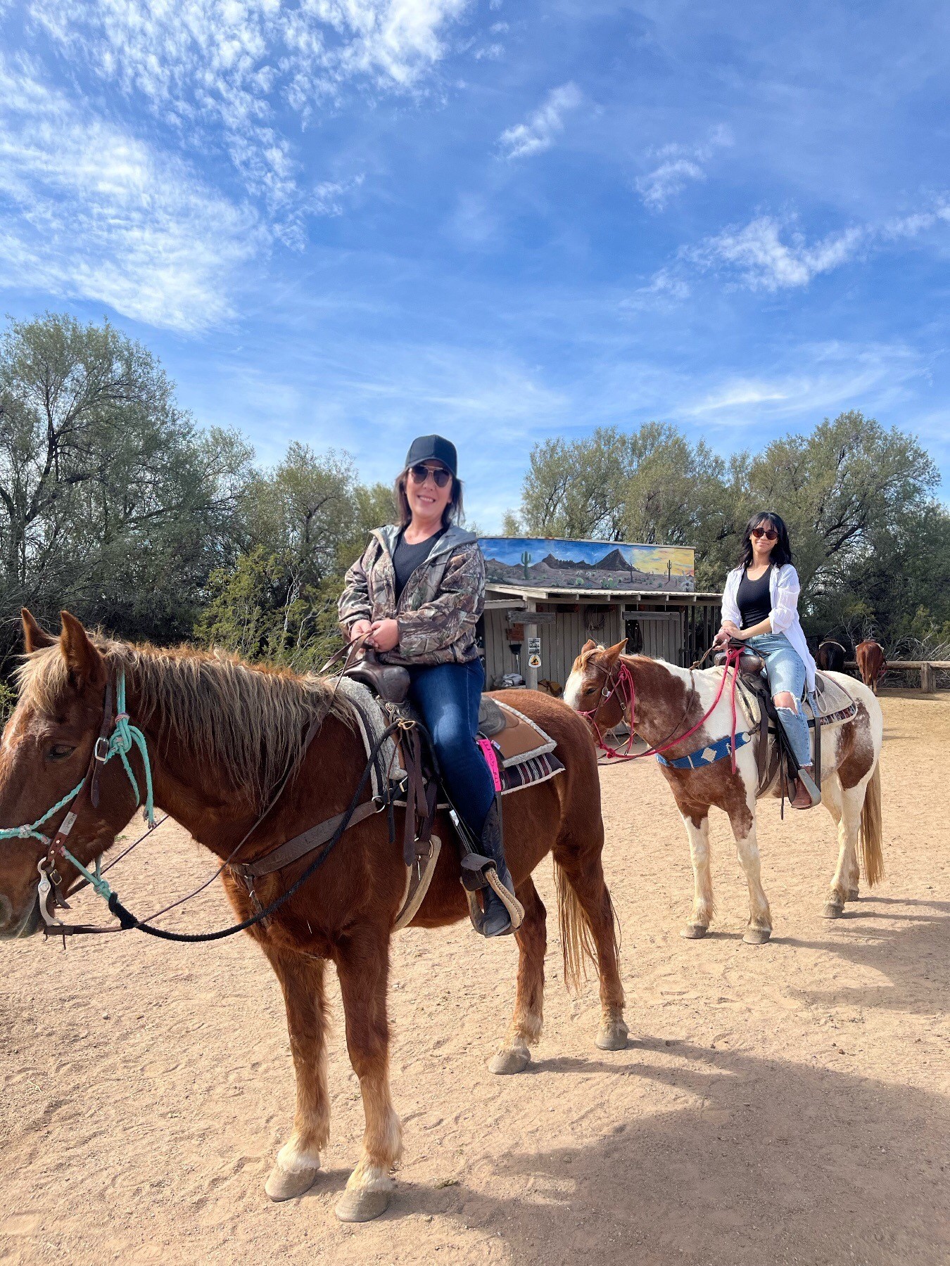 Two people horseback riding under a clear sky, with trees and a rustic building in the background.