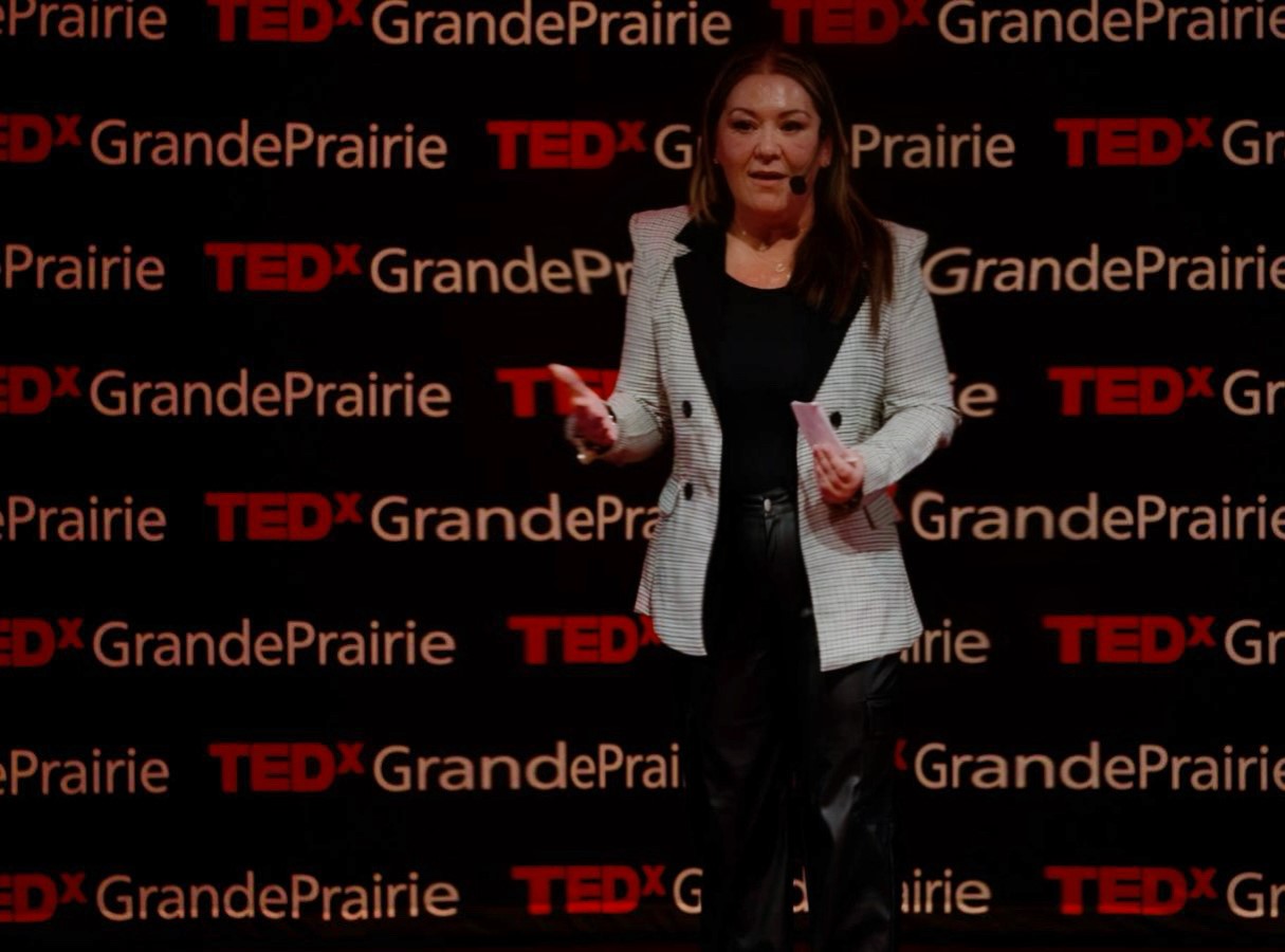 A person is giving a presentation on a TEDxGrandPrairie stage, standing in front of a black background with the event's logo visible. The speaker is dressed in a black blazer and pants, gesturing mid-speech.