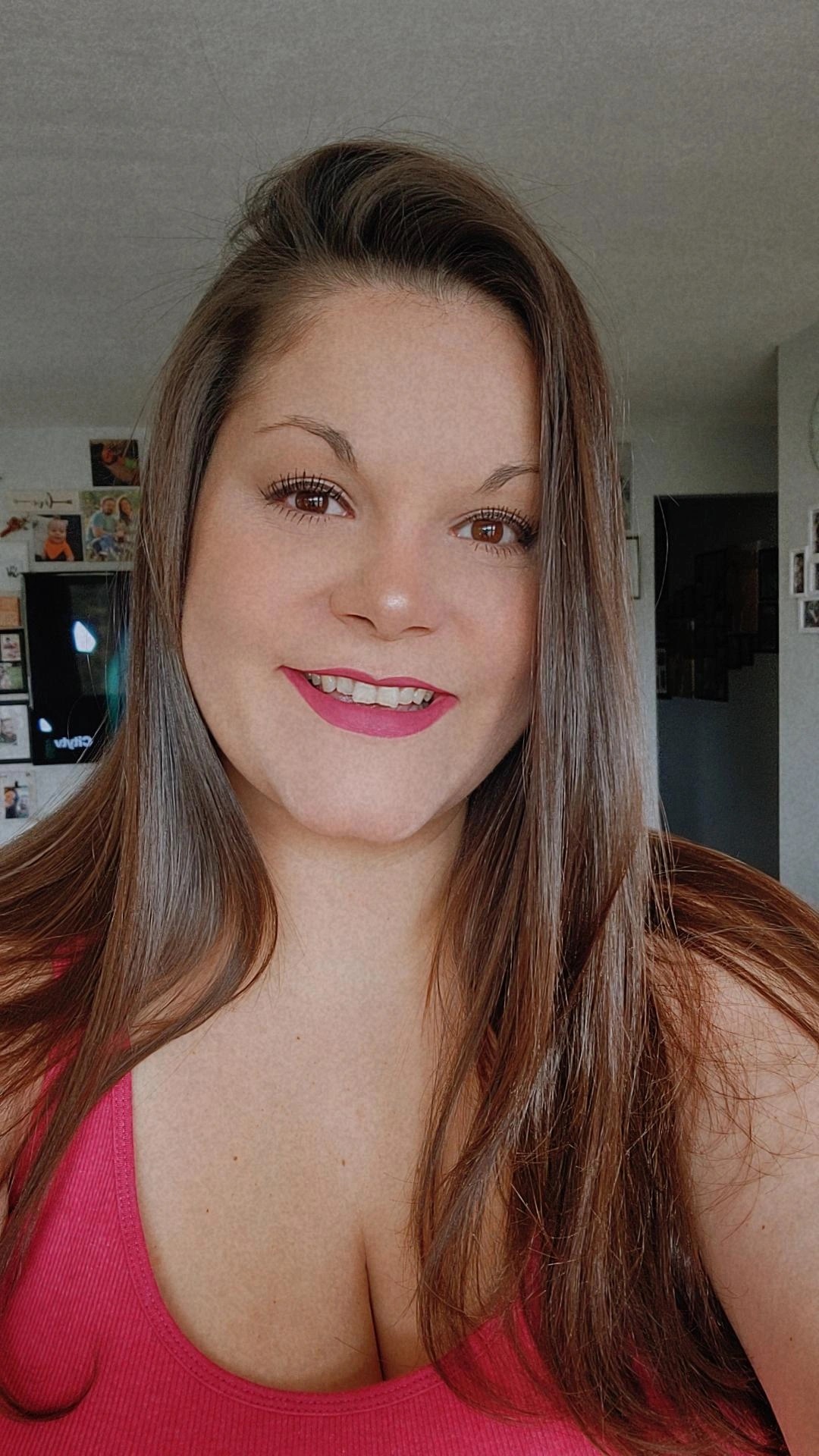 A selfie of a smiling woman with long brown hair and red lipstick wearing a red tank top