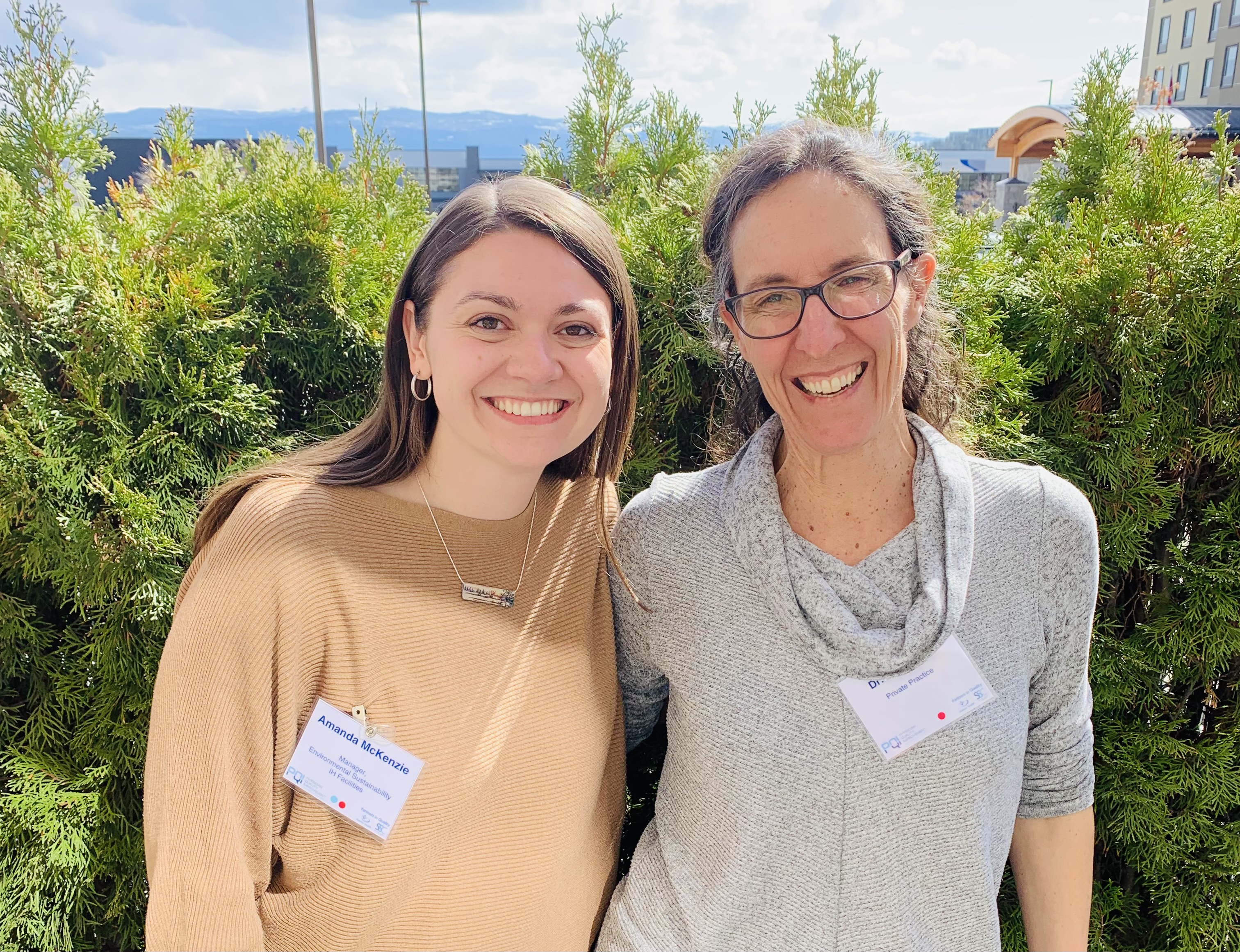 Two smiling people stand side by side with arms around each other, one with long dark hair in a beige sweater and one with glasses and curly grey hair in a grey sweater both standing in front of a hedge