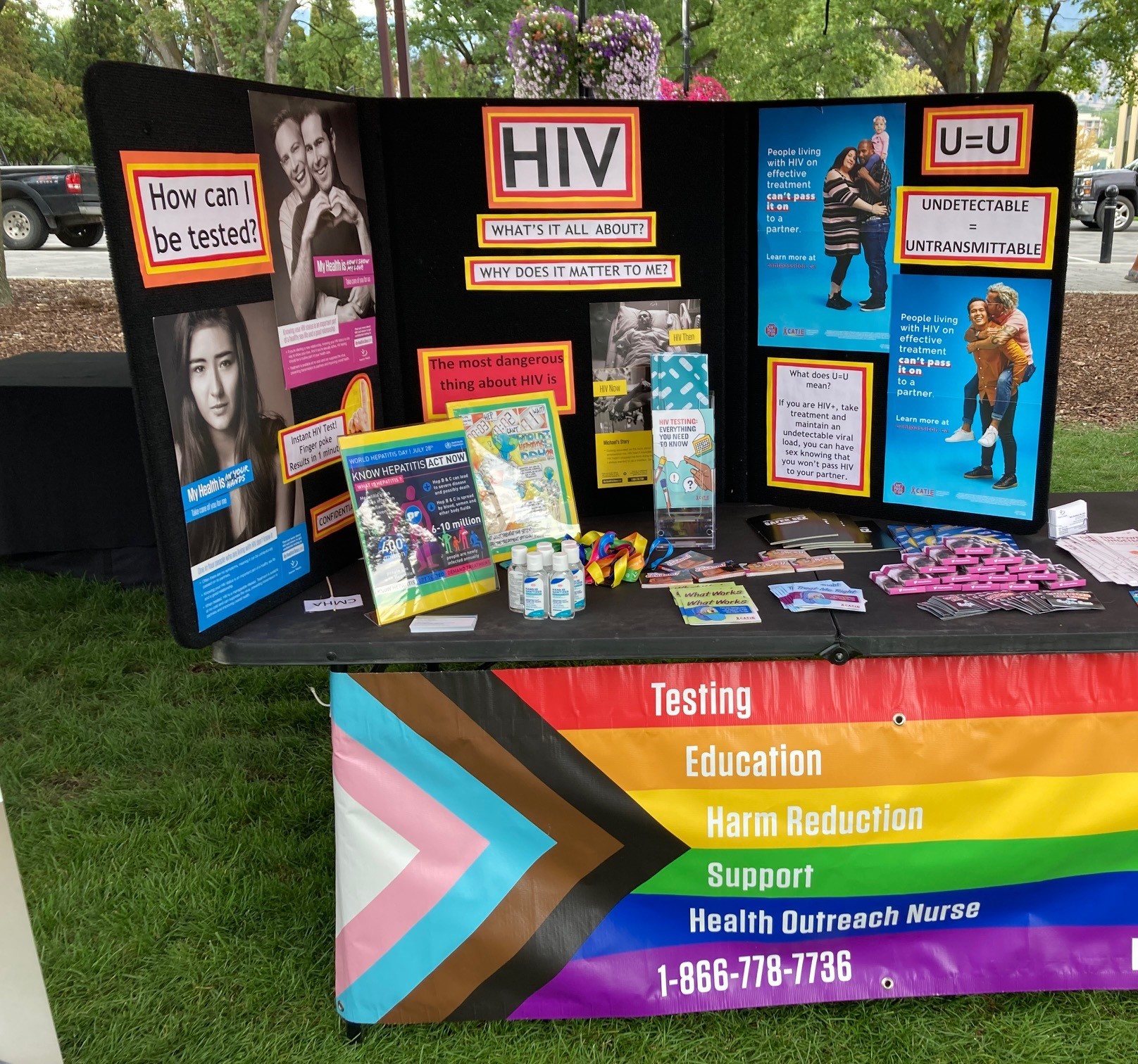  A health information booth featuring a colorful informational display on HIV awareness and prevention. Visible are educational pamphlets, posters, a rainbow table banner reading "Testing, Education, Harm Reduction, Support," and various promotional items.