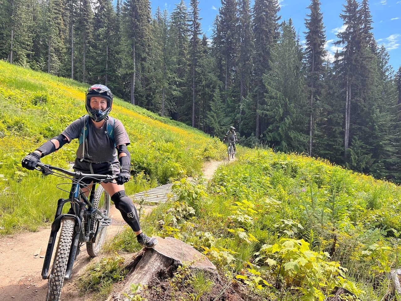 Woman sits on mountain bike, wearing knee pads, arm pads, gloves, and a full helmet. She is on a mountainside covered in greenery and yellow flowers, with evergreen trees in the background. Another cyclist is in the background further down the trail.