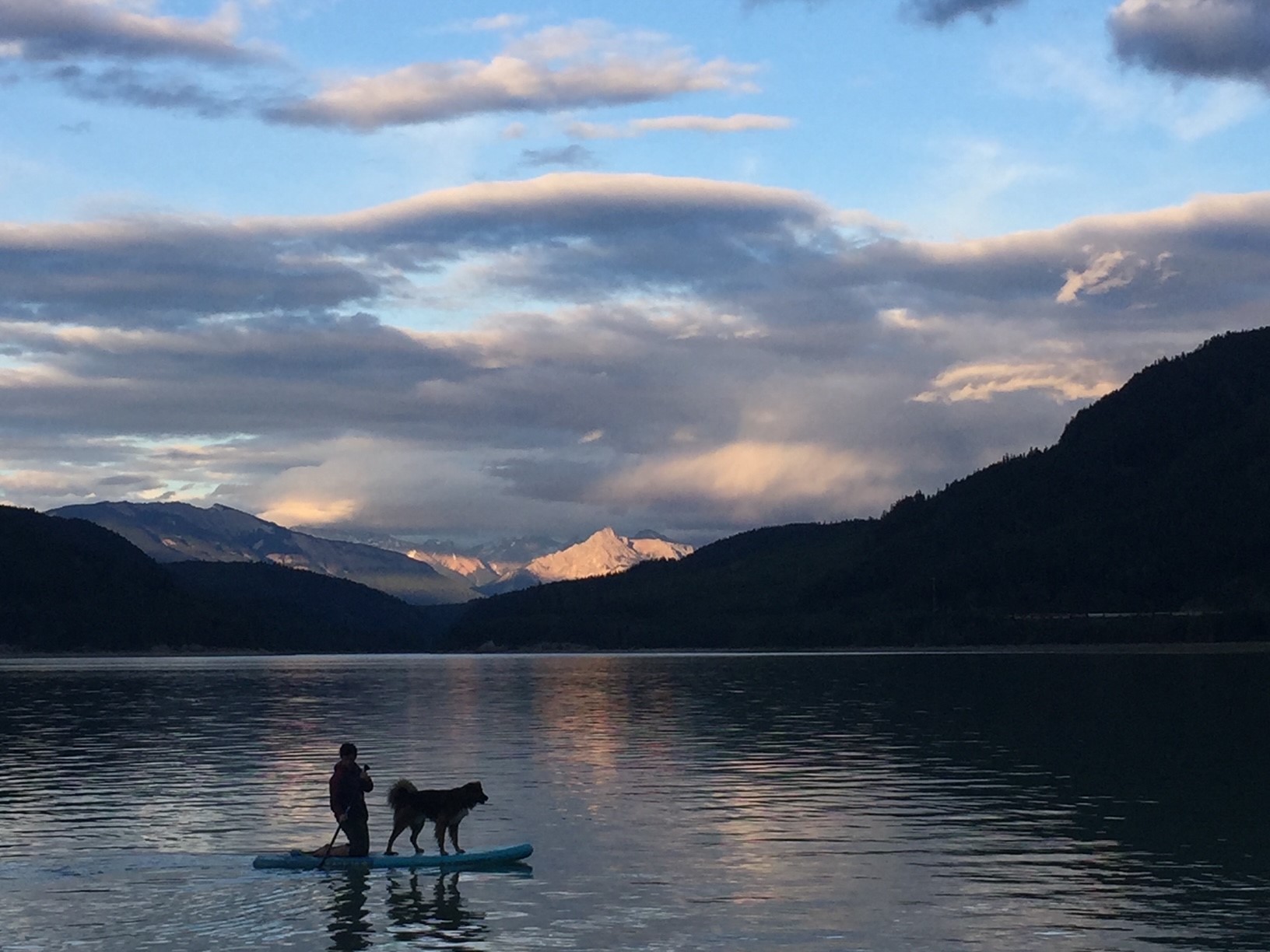 A lake with snow-capped mountains in the background at sunset. On the lake is the silhouette of a person and a dog on a paddleboard. 