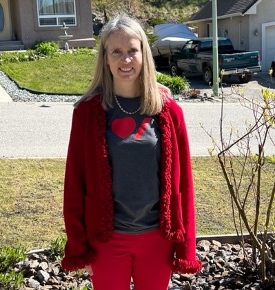 A smiling woman with blond hair wearing a red sweater, red pants and a grey t-shirt with a heart stands on a street in front of homes and a black pickup truck in the background