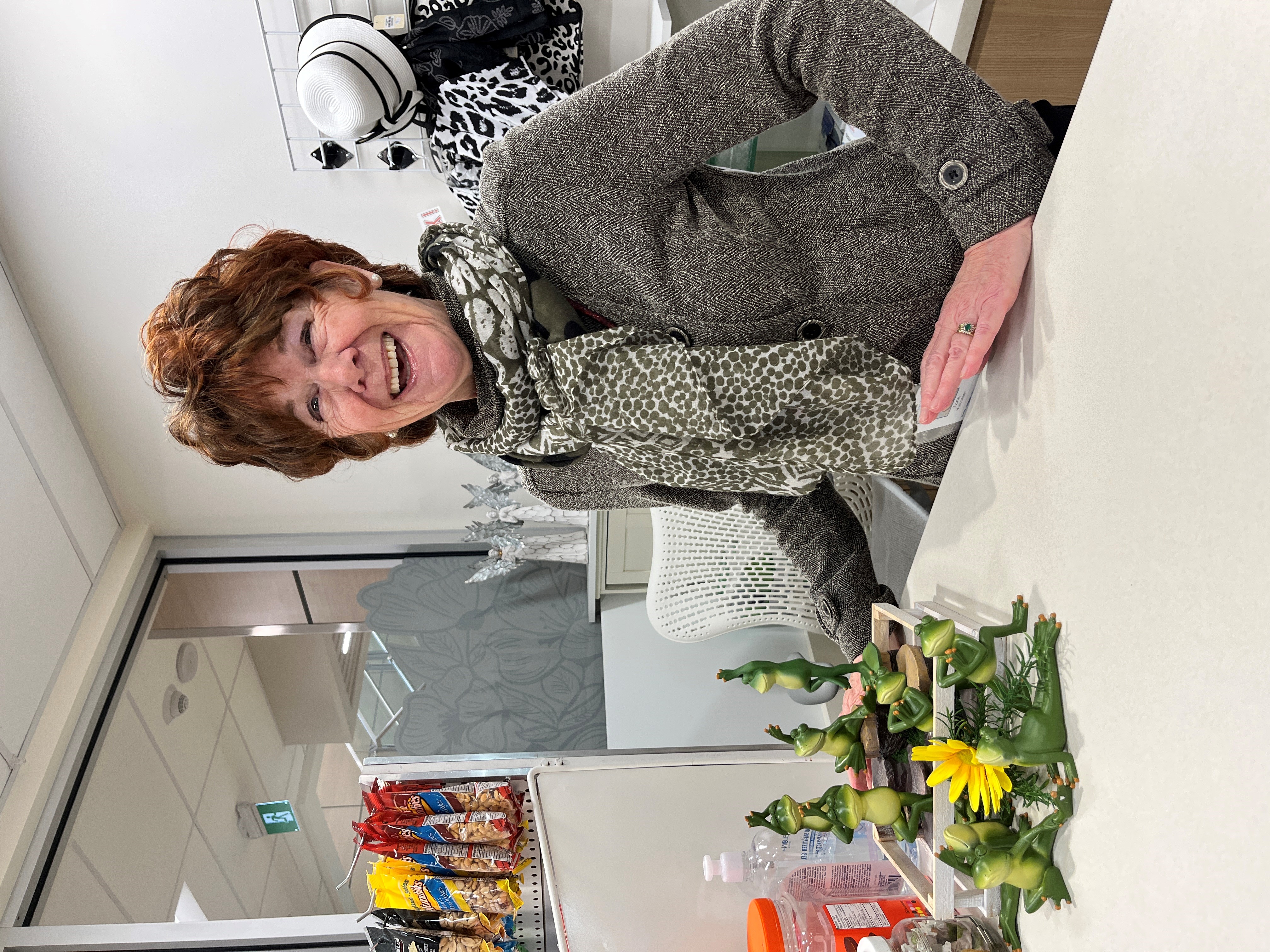 A person laughing and wearing a grey herringbone jacket and scarf stands behind a counter with a display of glass frog ornaments