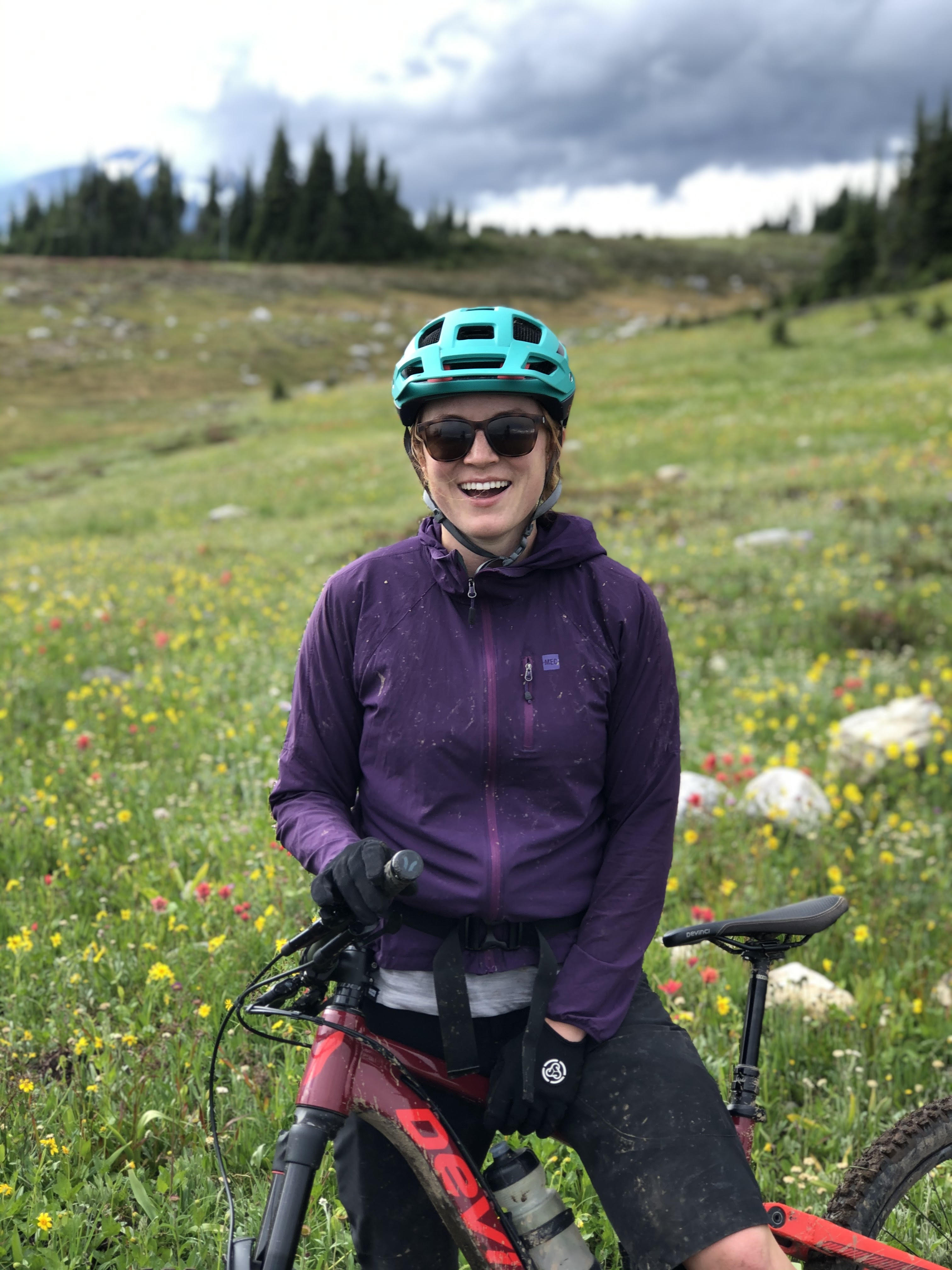 A person in a teal bike helmet, sunglasses and a purple jacket rests on a grey and red mountain bike in a field of flowers and trees