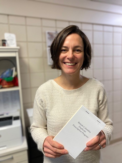 A smiling person wearing a white sweater standing in front of a tiled wall holds a prescription pad