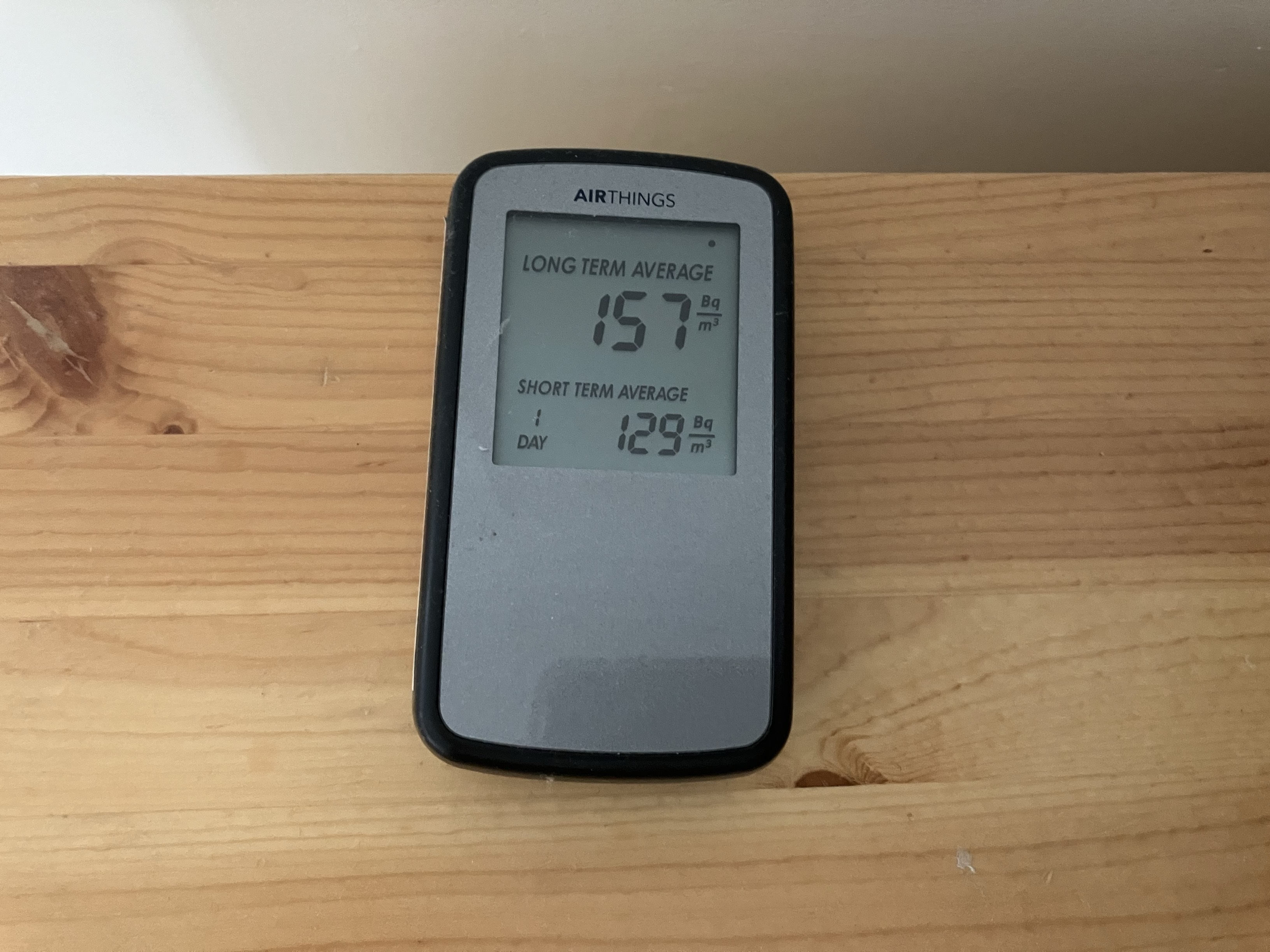 AirThings radon detector displaying long term average at 157 Bequerels per metre cubed and short term average at 129 Bequerels per metre cubed on a wooden shelf.