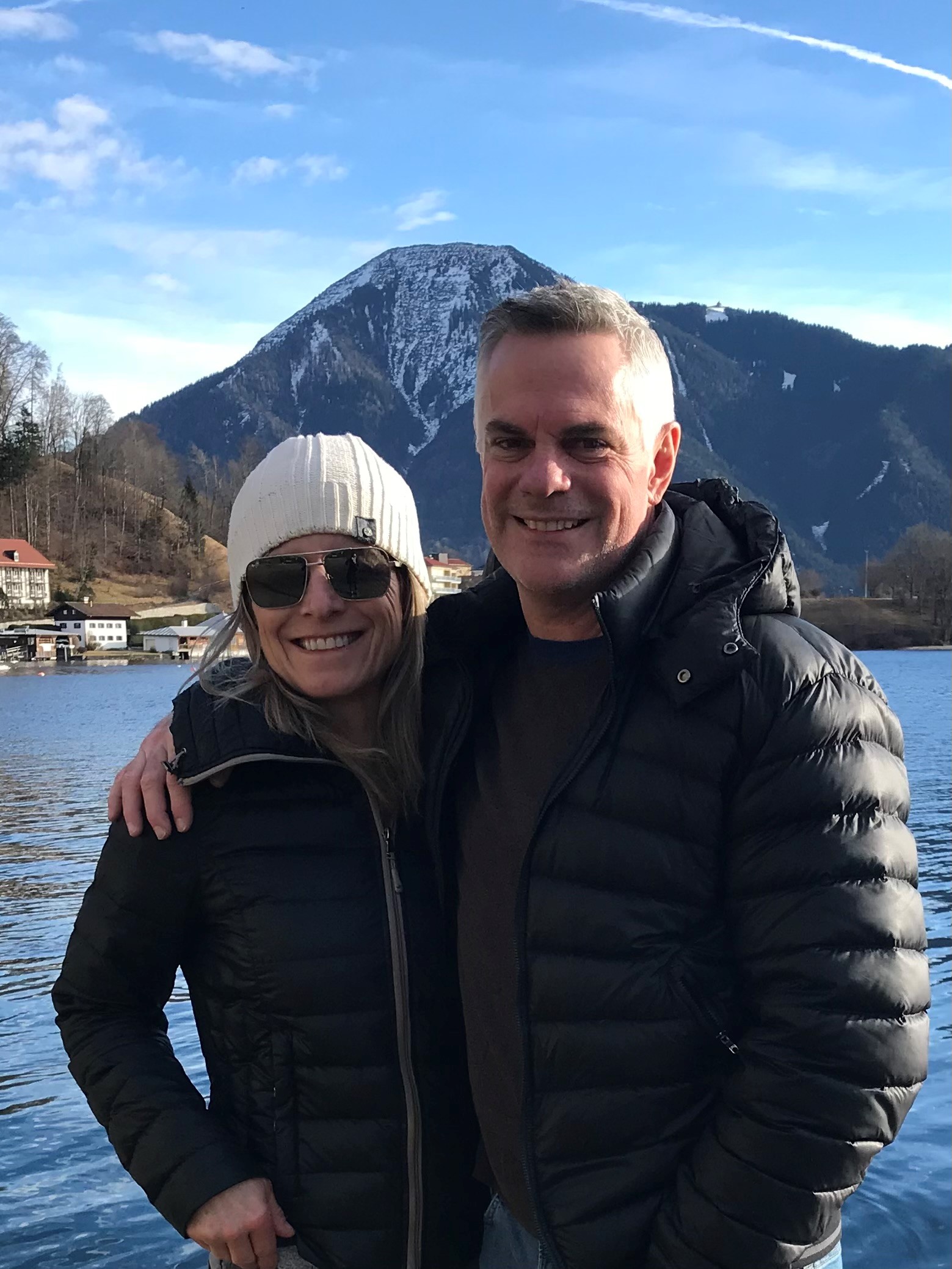 A man and woman wearing winter gear pose for a picture together outside with water and mountains in the background.