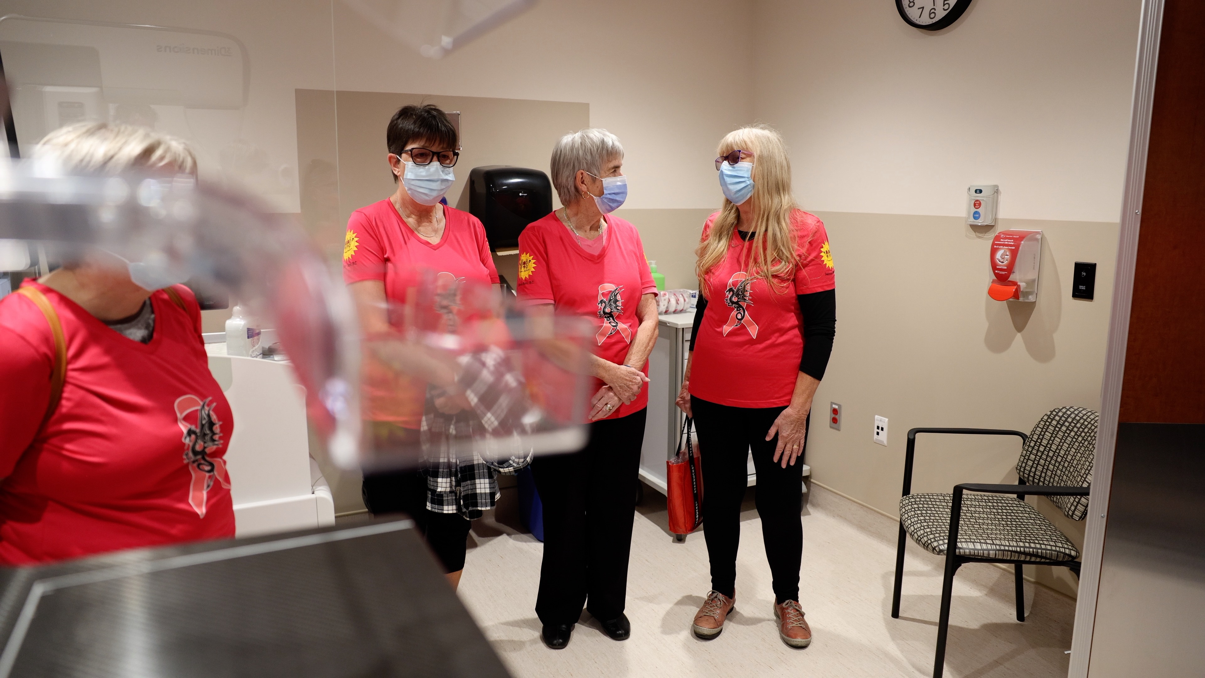 Three people wearing pink shirts and masks stand and talk inside.