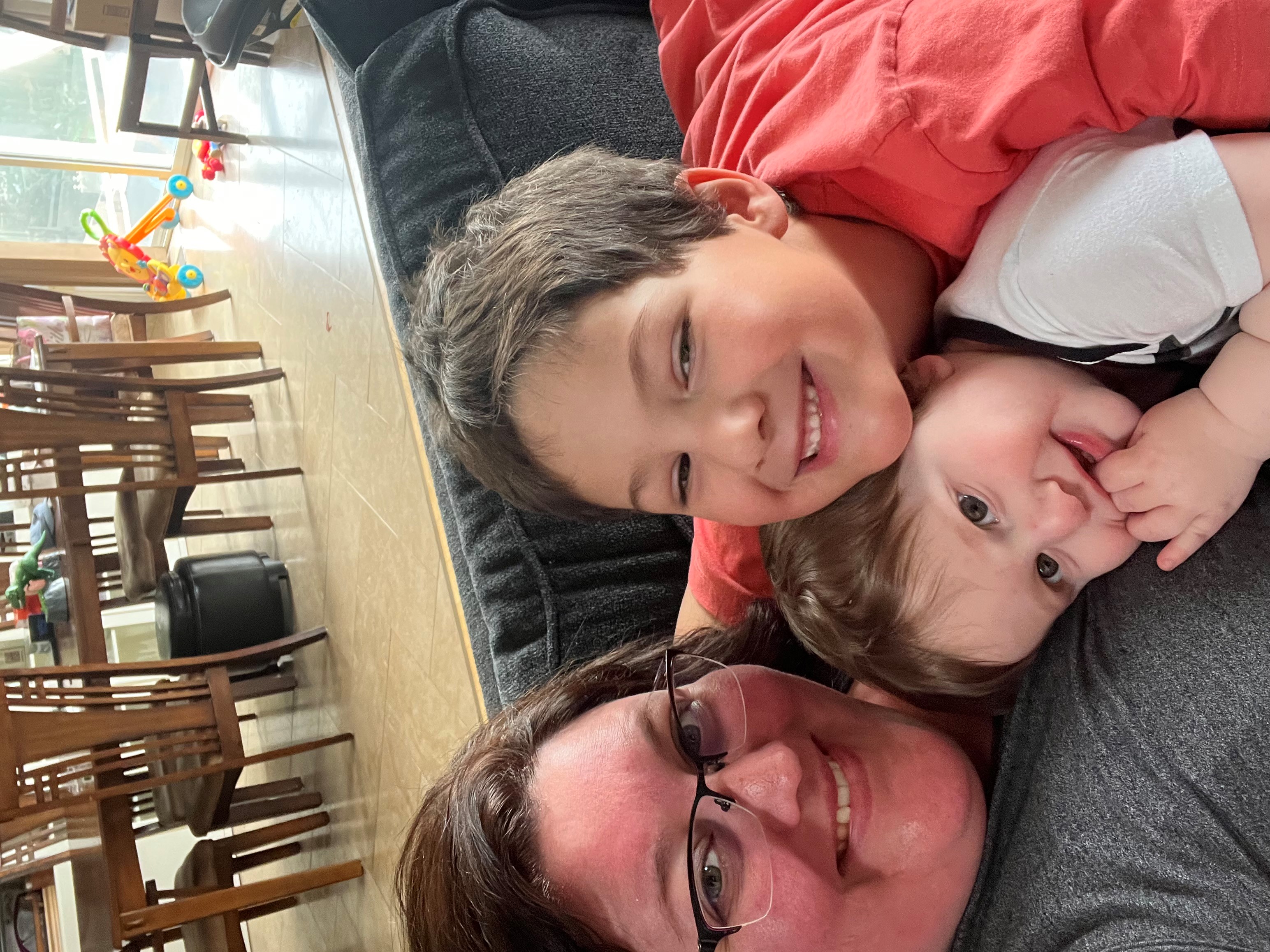 A smiling woman with glasses and grey shirt sits on a grey couch cuddling two young boys