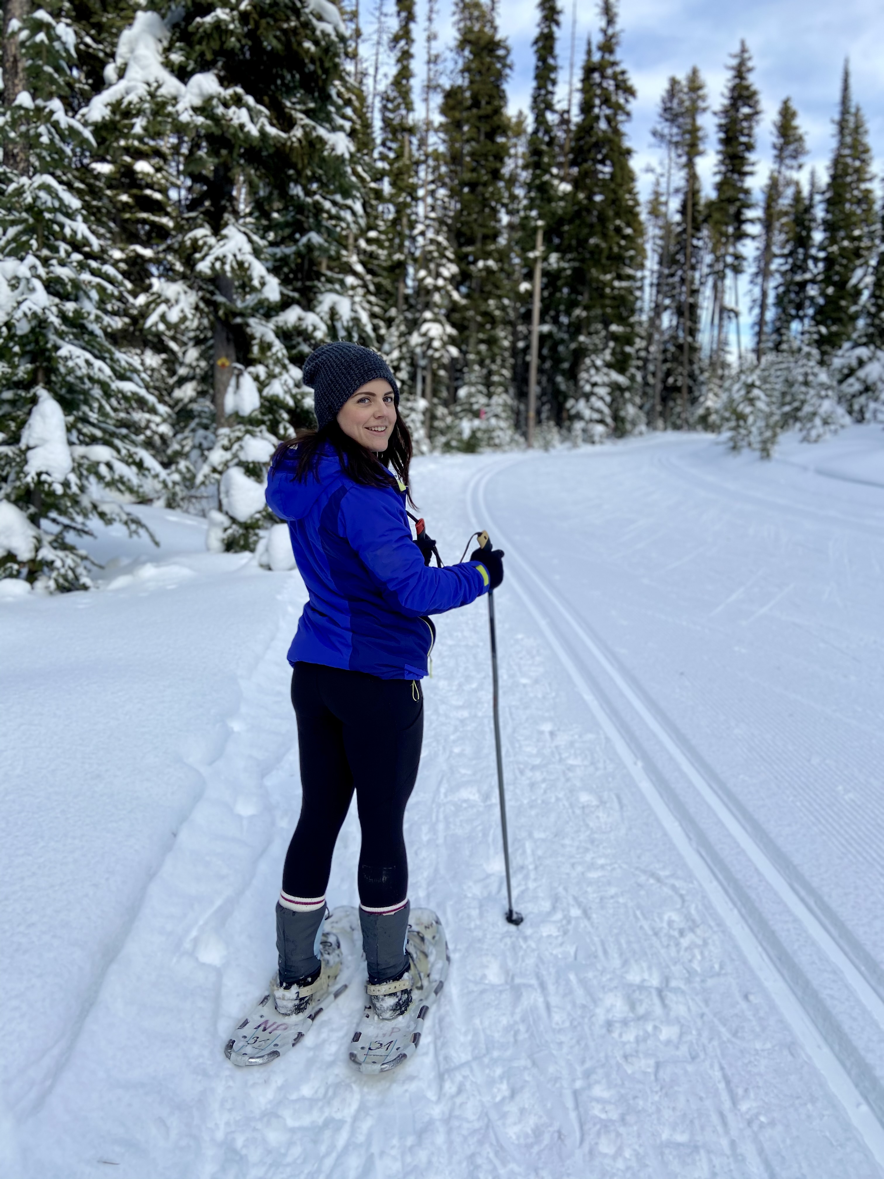 A smiling woman in a toque and bright blue snow jacket and black pants holds poles while snowshoeing on a snowy path in a forest