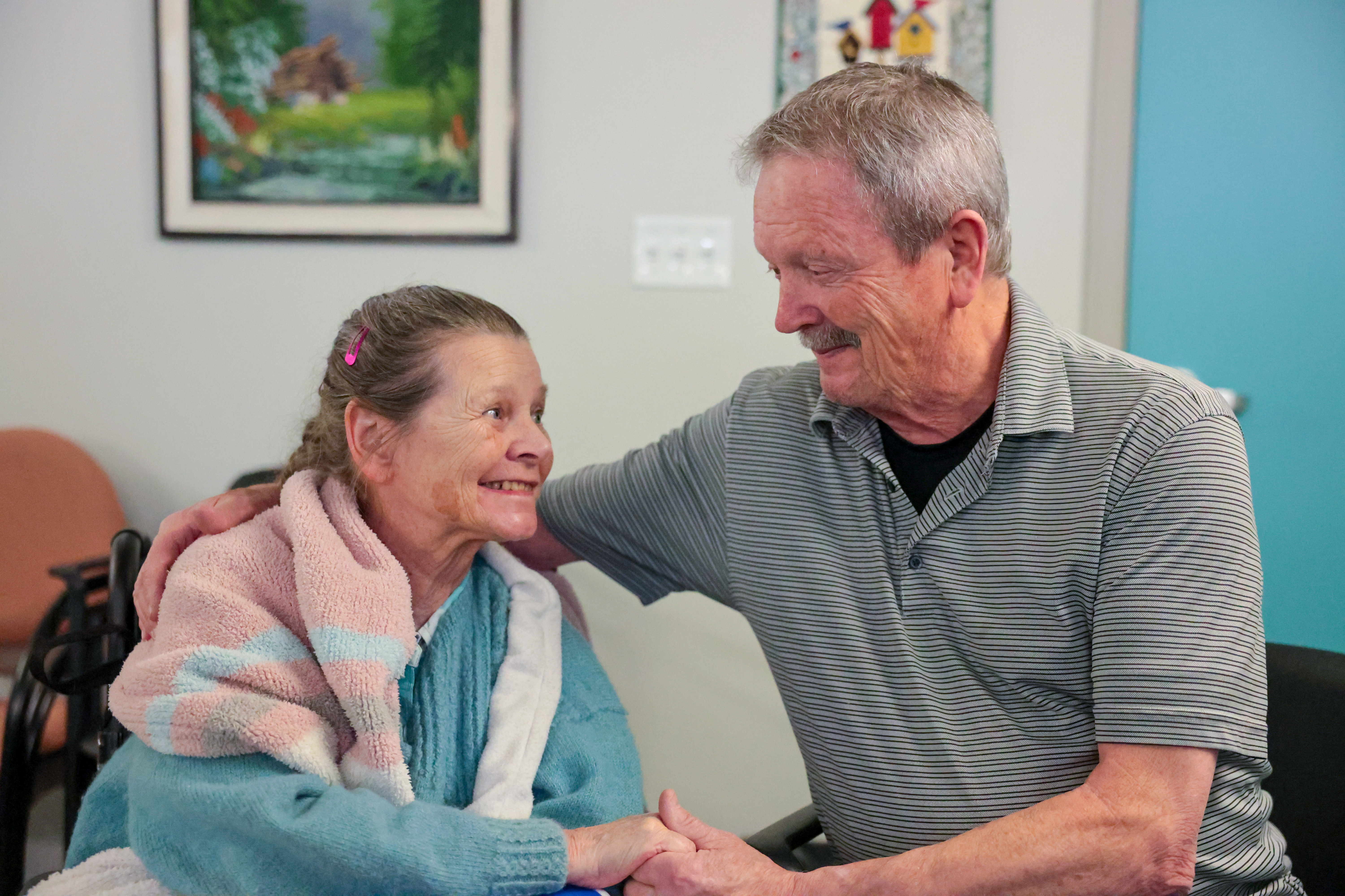 A woman with a long grey braid, pink shawl and teal sweater smiles and holds hands with a man with grey hair and mustache in a grey polo shirt