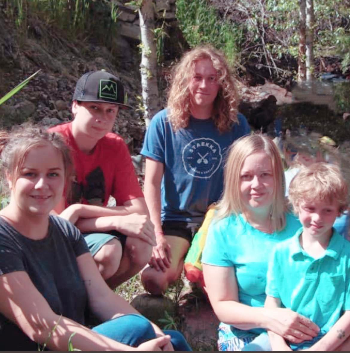 A family of five sit together outside. Two young men, a young boy, a young woman, and a mother holding the young boy smile to camera. 