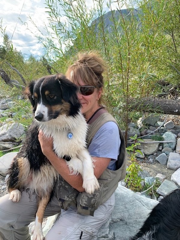 A person outdoors holding a black and white dog, surrounded by rocky terrain and greenery.