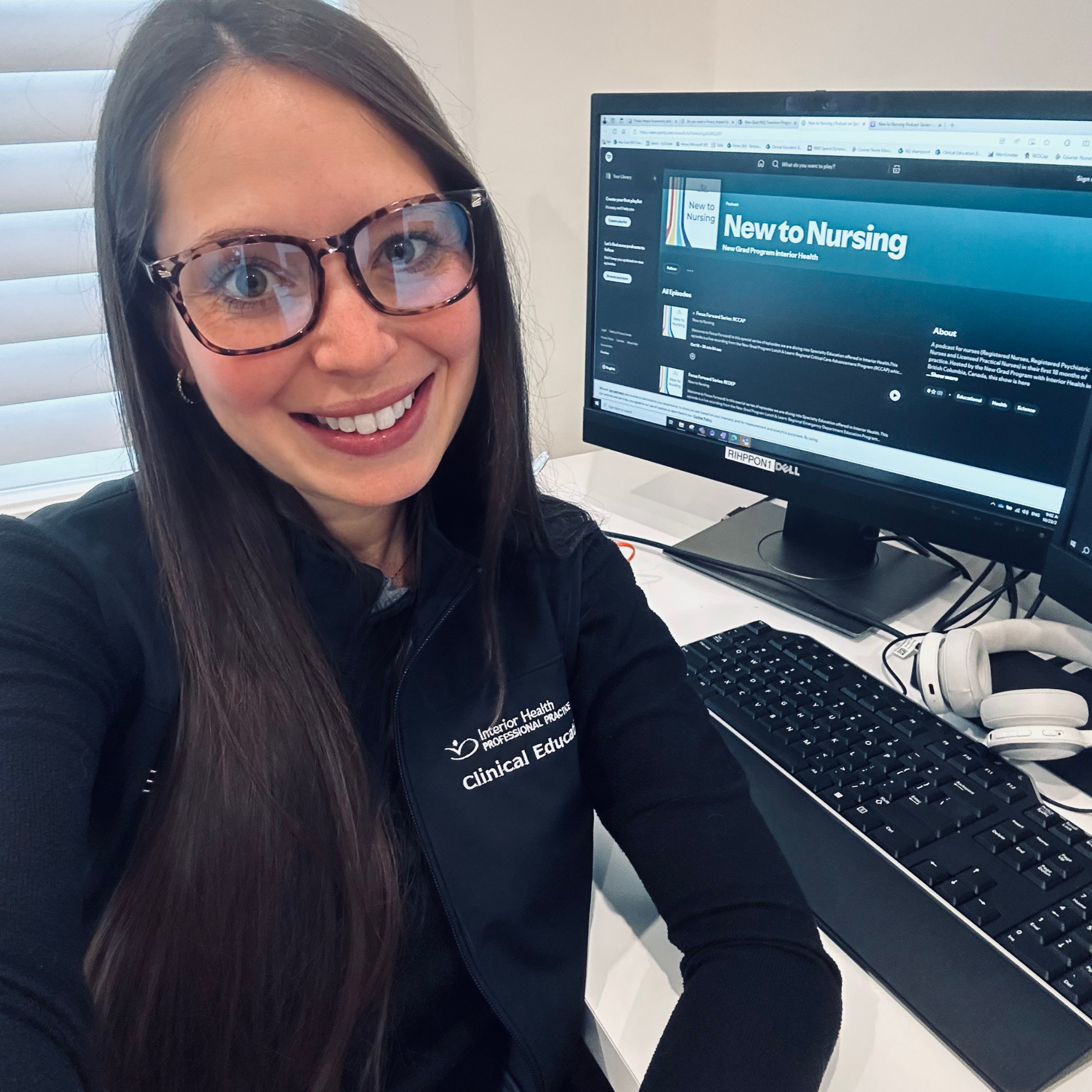 Person smiling at the camera, wearing glasses and a 'Clinical Educator' uniform, seated at a desk with a computer showing a 'New to Nursing' webpage.