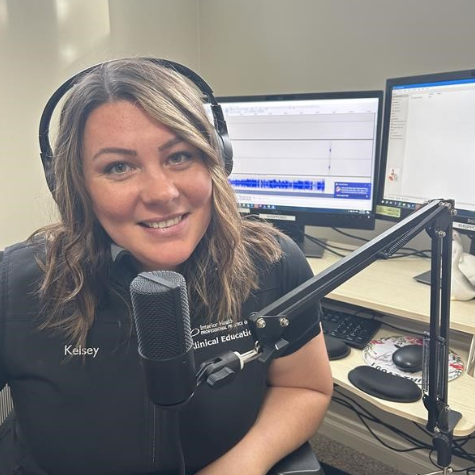 Person named Kelsey in a clinical research uniform sitting at a desk with headphones on, speaking into a microphone, with two computer screens displaying data charts in the background.