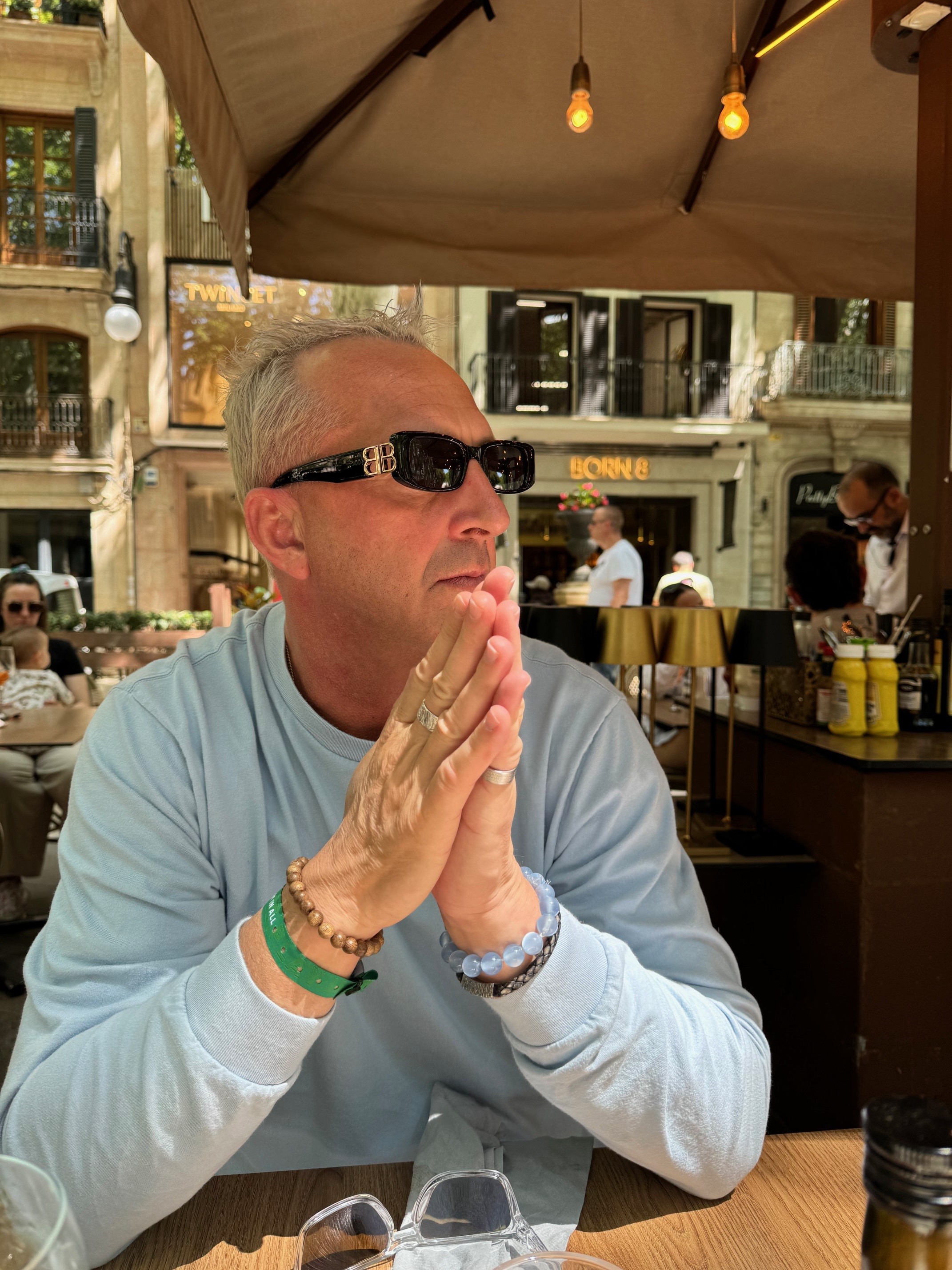 Person sitting at a café table with hands pressed together, wearing sunglasses, in front of a street view with visible café signs.