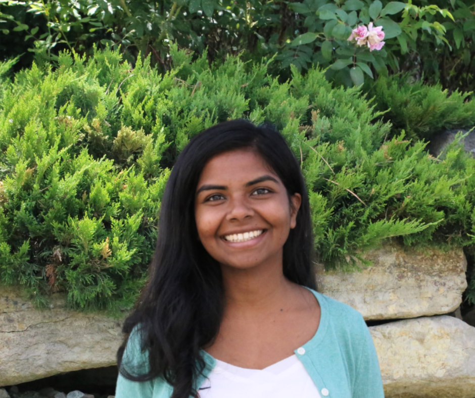 A smiling person with long black hair wearing a teal sweater and white t-shirt stands in front of shrubs and a rock wall