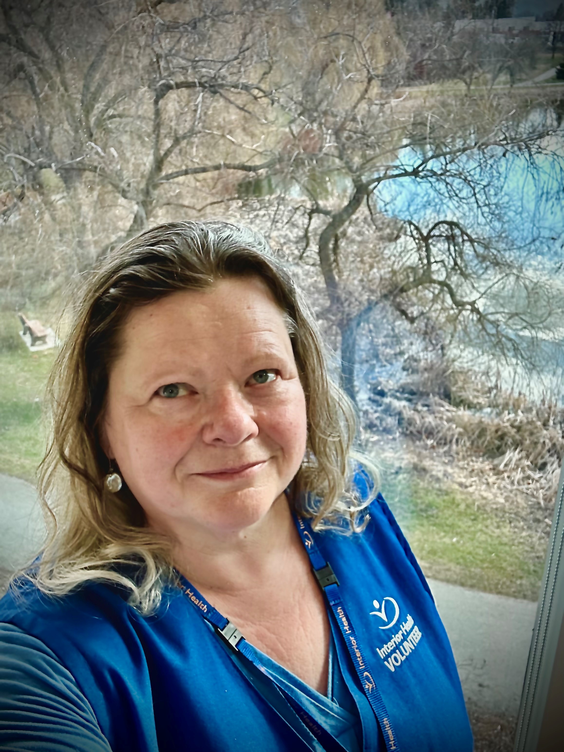 A person with curly medium blonde hair wearing a blue Interior Health vest stands in front of trees without leaves