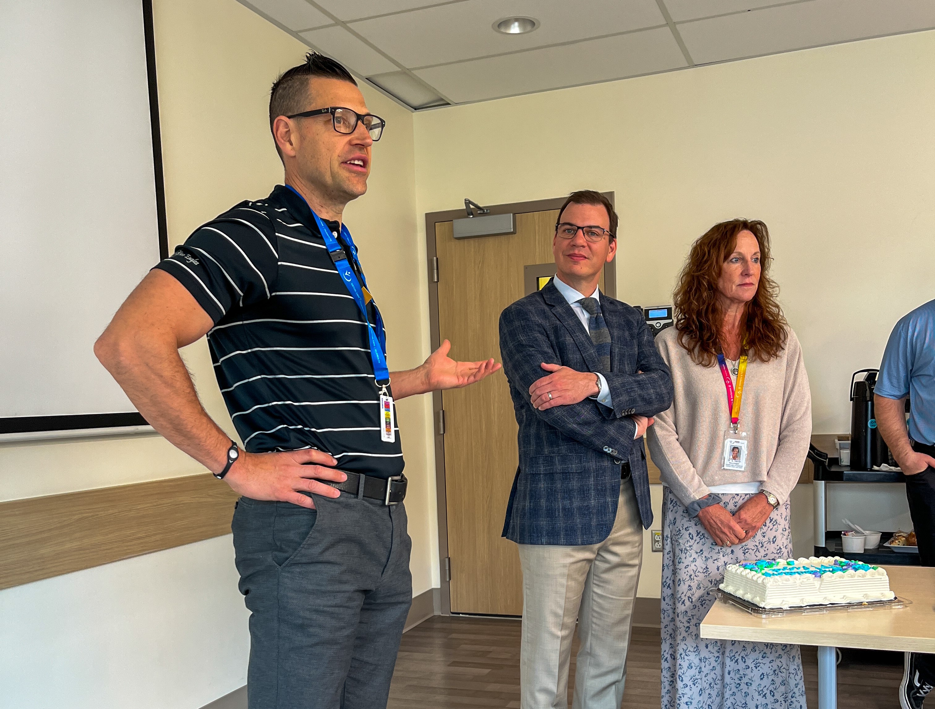 Person standing and speaking in a room with three attentive listeners; all individuals display hospital ID badges. A cake on a rolling table is also visible.