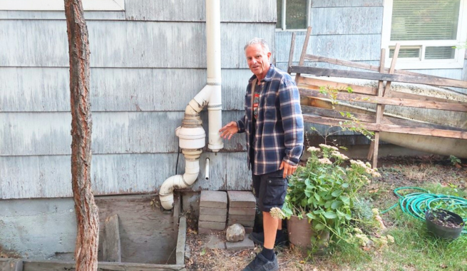 A man in a checkered jacket and black shorts and sandals points at a white vertical pipe adjacent to a blue shingled house