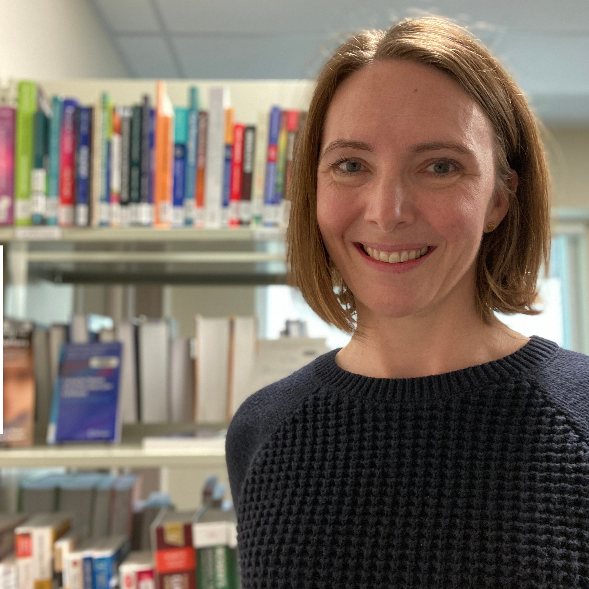 A smiling person with medium length brown hair and a black top standing in front of a bookshelf in a library setting.