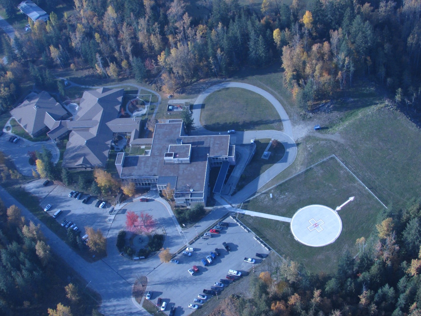 An overhead view of a hospital with several buildings and a helicopter pad surrounded by trees with green and yellow leaves