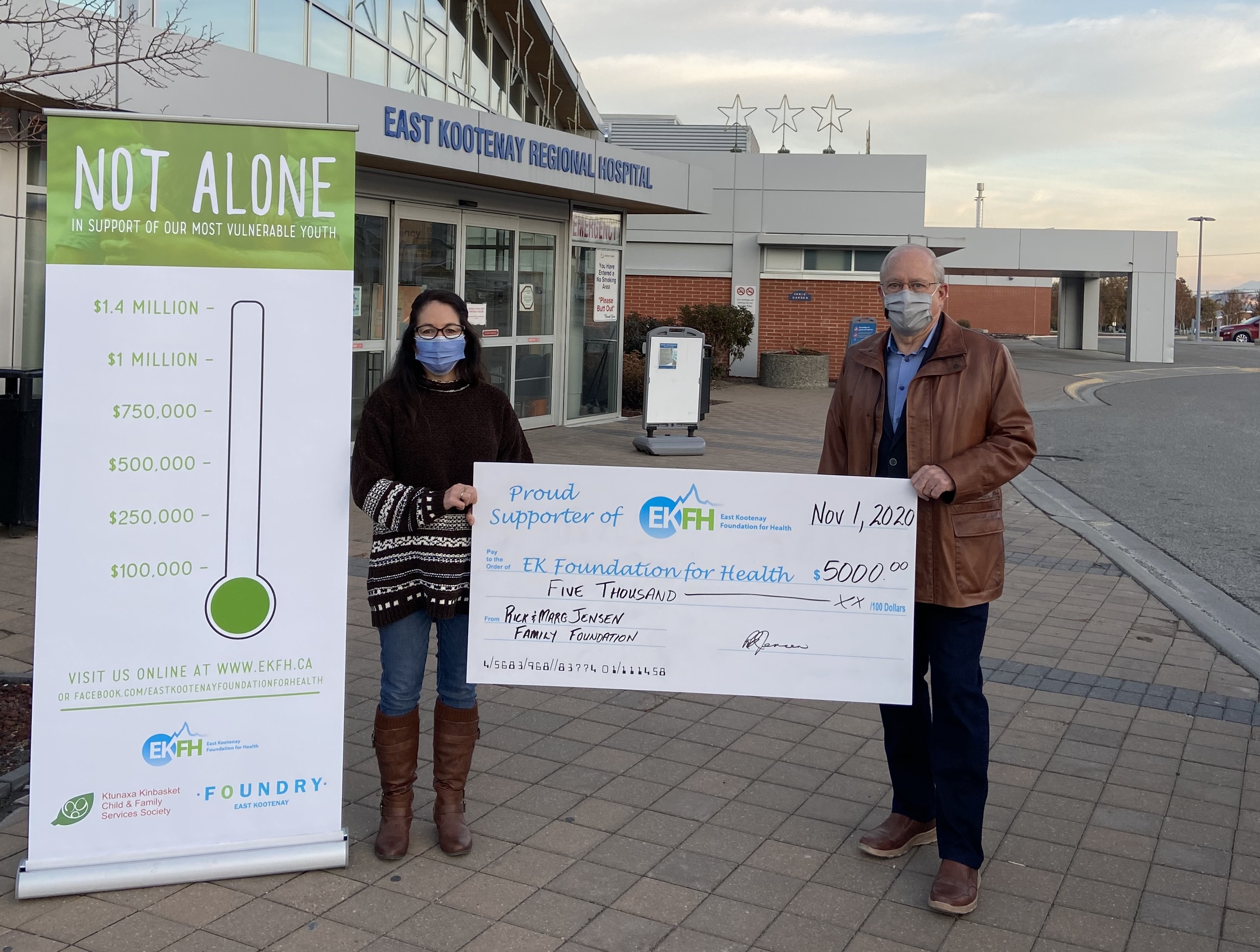 A woman with long dark hair and wearing a face mask, long black sweater, blue jeans and boots holds a large cheque with a man with grey hair, glasses, a face mask, brown leather jacket and black pants in front of a hospital and sign that says Not Alone In support of our most vulnerable youth