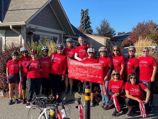 A group of 14 people wearing red t-shirts and bike helmets hold a sign that says women's heart health in front of a building on a sunny day