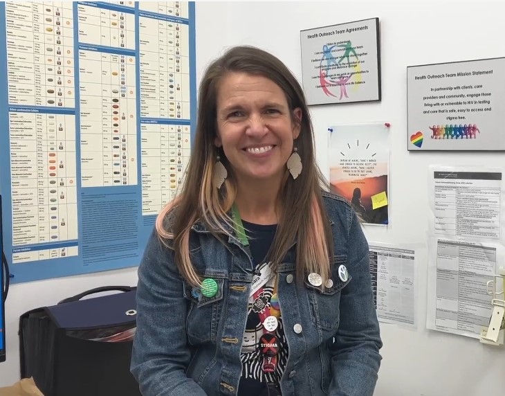 A smiling person wearing a jean jacket and pins with leaf earrings, sitting in a medical office with posters in the background.