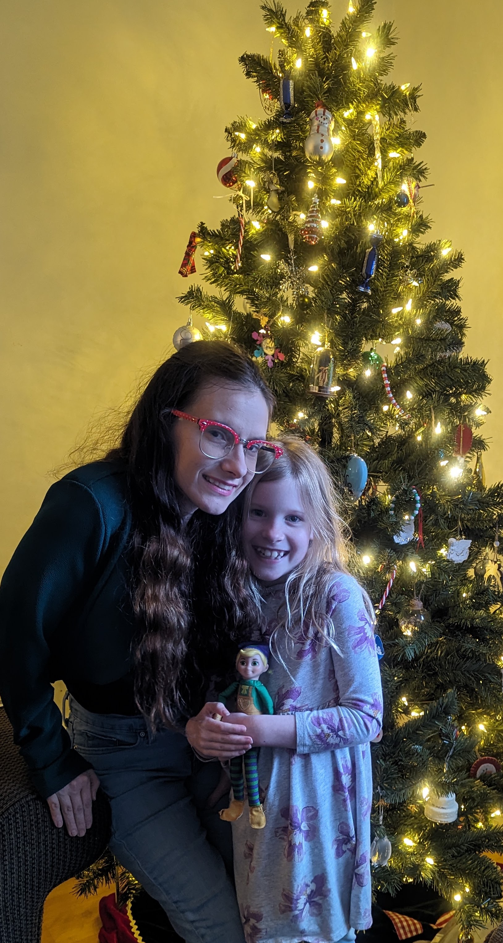 A woman and a child smile and pose together in front of a Christmas Tree