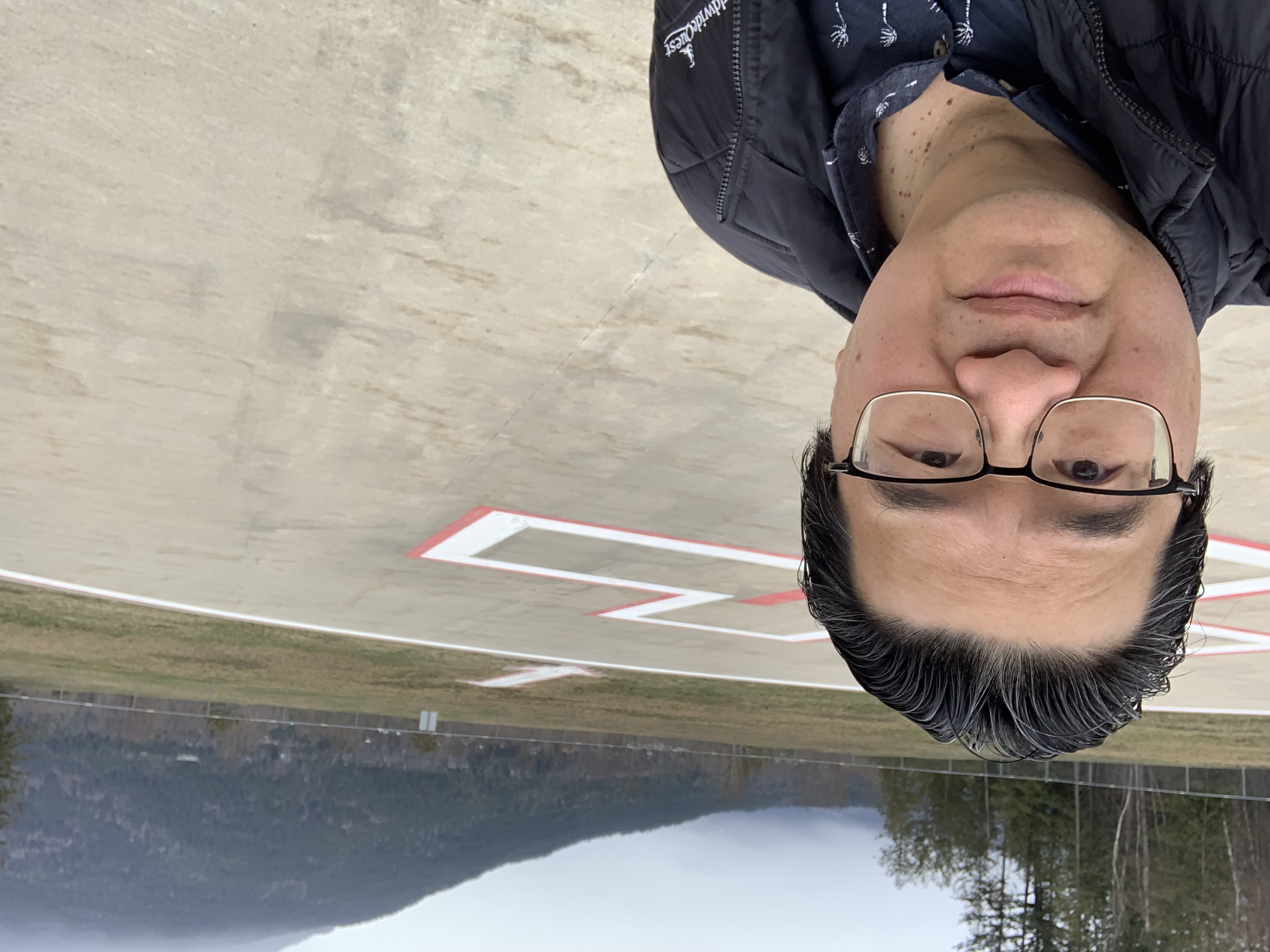 A man with combed back black hair and silver glasses and wearing a black jacket takes a selfie in a parking lot