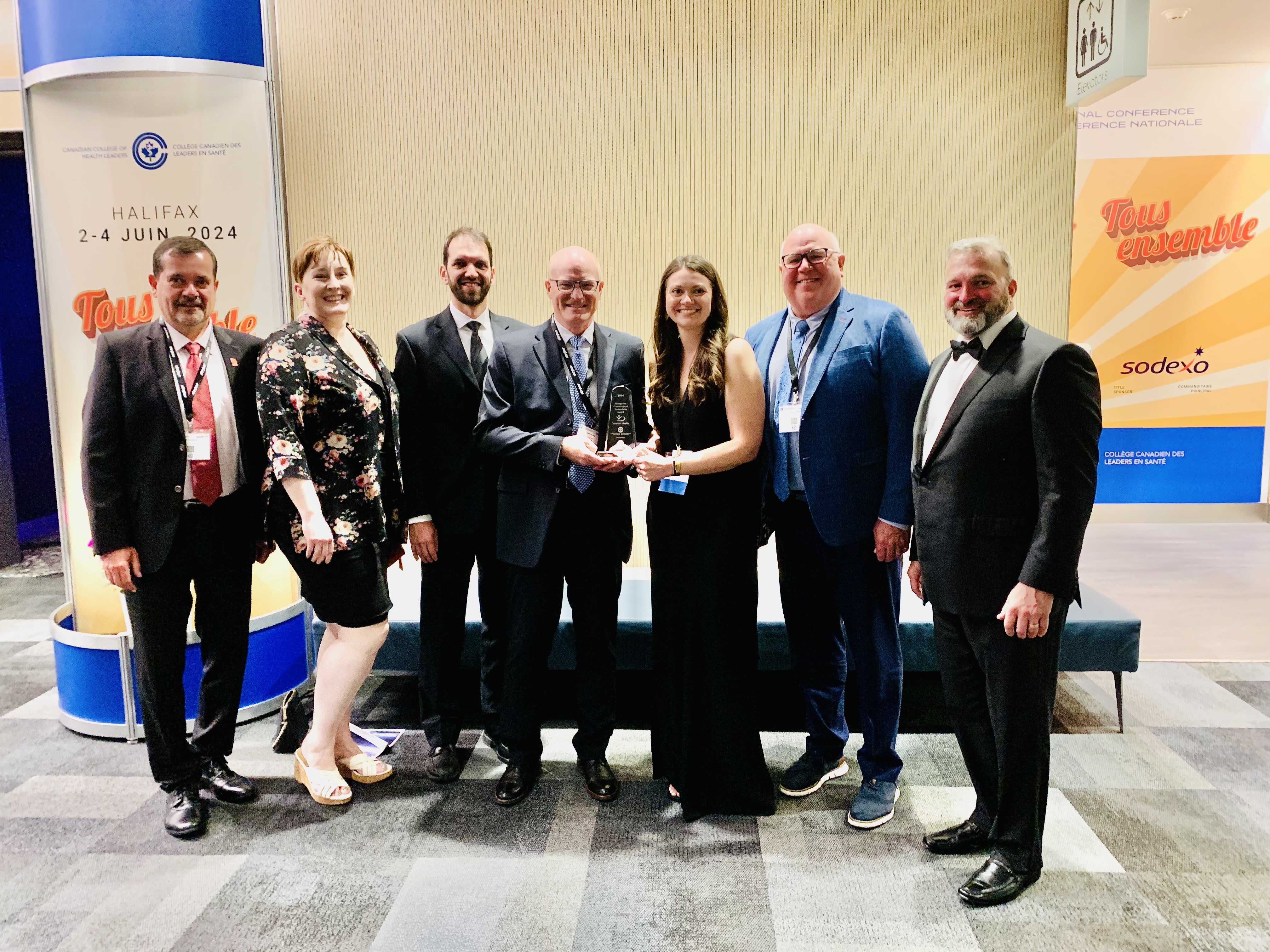 Group of six individuals in formal attire holding a trophy at a conference, standing in front of banners that advertise future events and sponsors including "Halifax 2-4 June 2024" and "Sodexo."