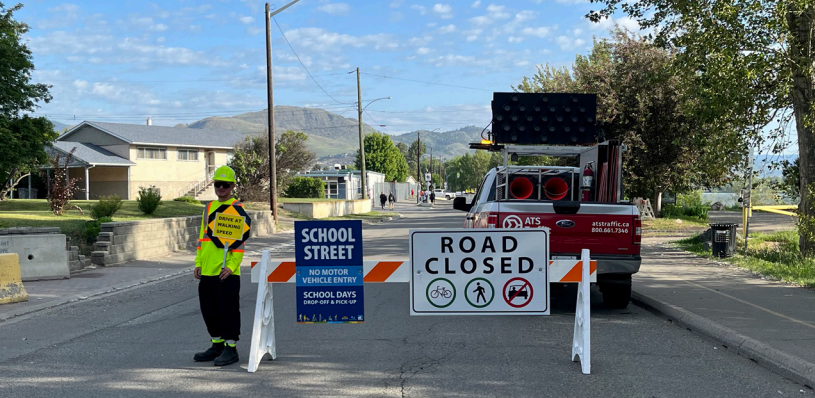 A traffic officer in a reflective vest stands next to a "Road Closed&quot; sign and barriers on a street, with a digital message board and a vehicle in the background. Signs indicate &quot;School Street No Motor Vehicle Entry" with additional symbols for no cars, bikes, or trucks.