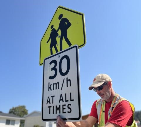 An individual in a reflective vest installs a road sign indicating a speed limit of 30 km/h, featuring an overhead caution graphic of an adult and child crossing.